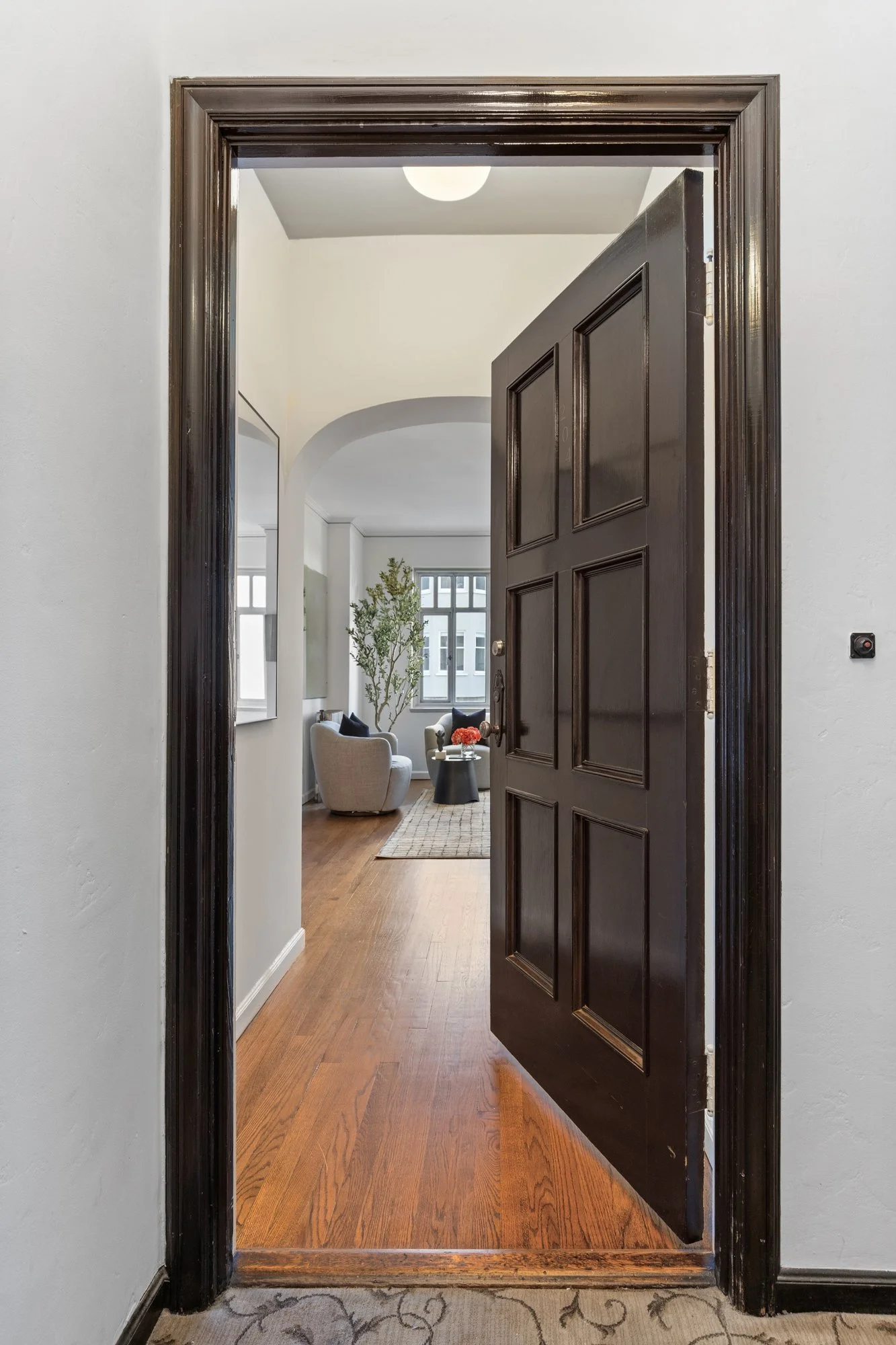 View through a dark wooden door into a living room with white walls, hardwood floors, and large windows. The room has a cozy seating area with a beige armchair, a small black side table with a vase of orange flowers, a potted plant, and abstract artw