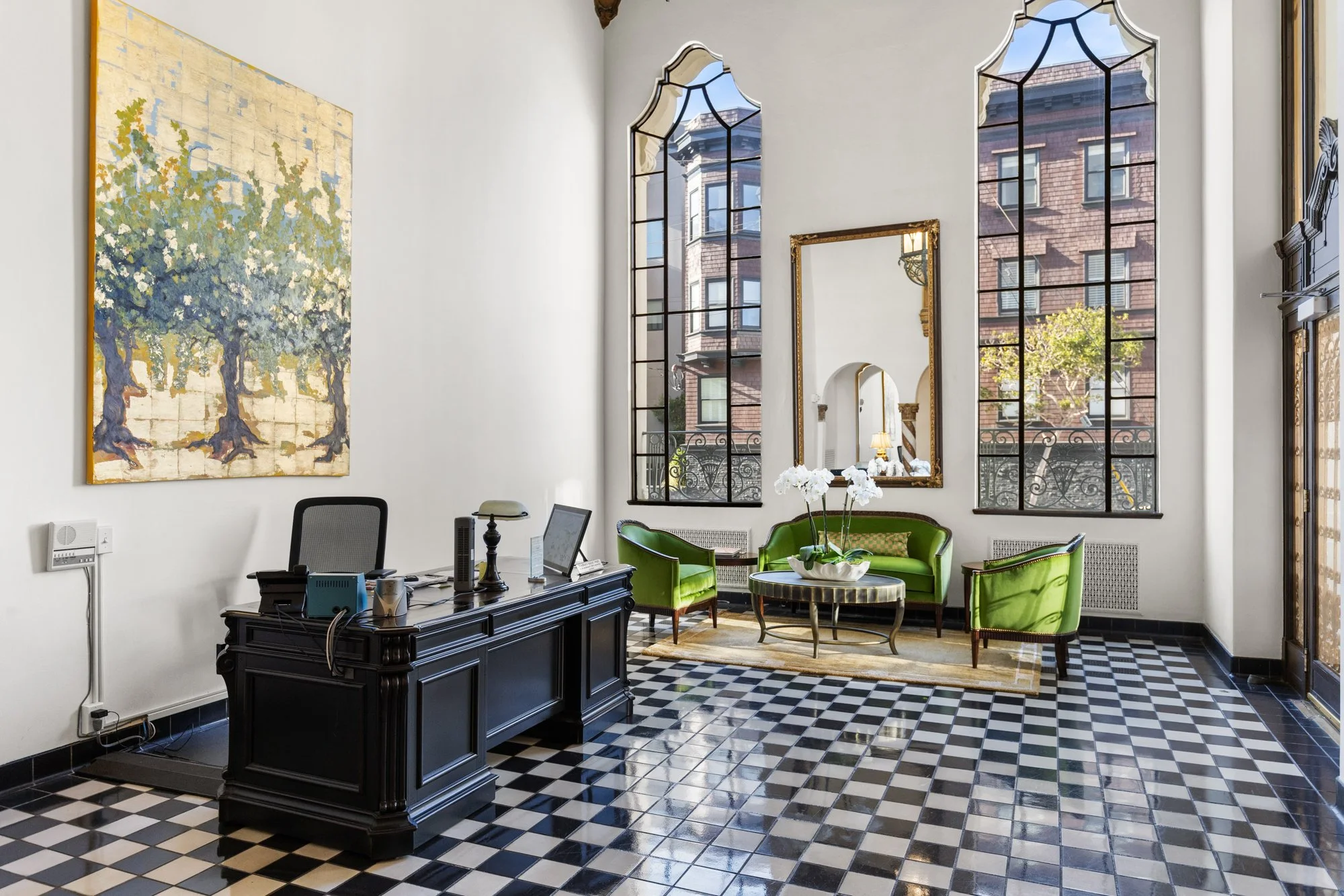 Elegant office space with large arched windows, a black desk with office equipment, a vintage mirror, a green settee and chairs, a round coffee table with white orchids, and artwork on the walls.