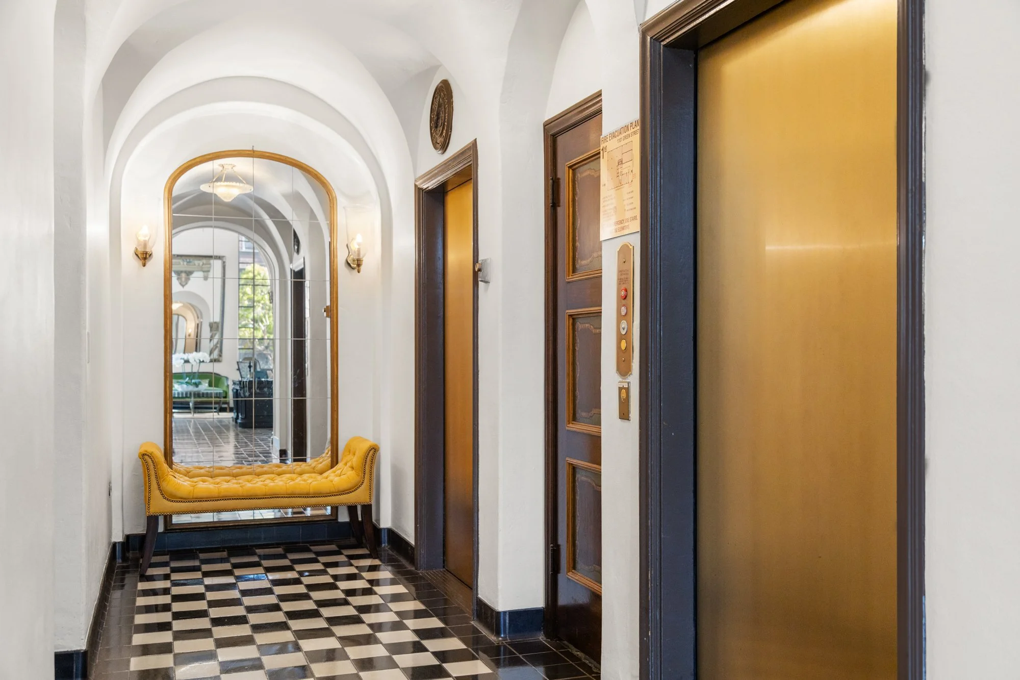 A hallway with black and white checkered tile floor, white curved ceilings, a large mirror, a mustard yellow tufted bench, and two elevator doors.