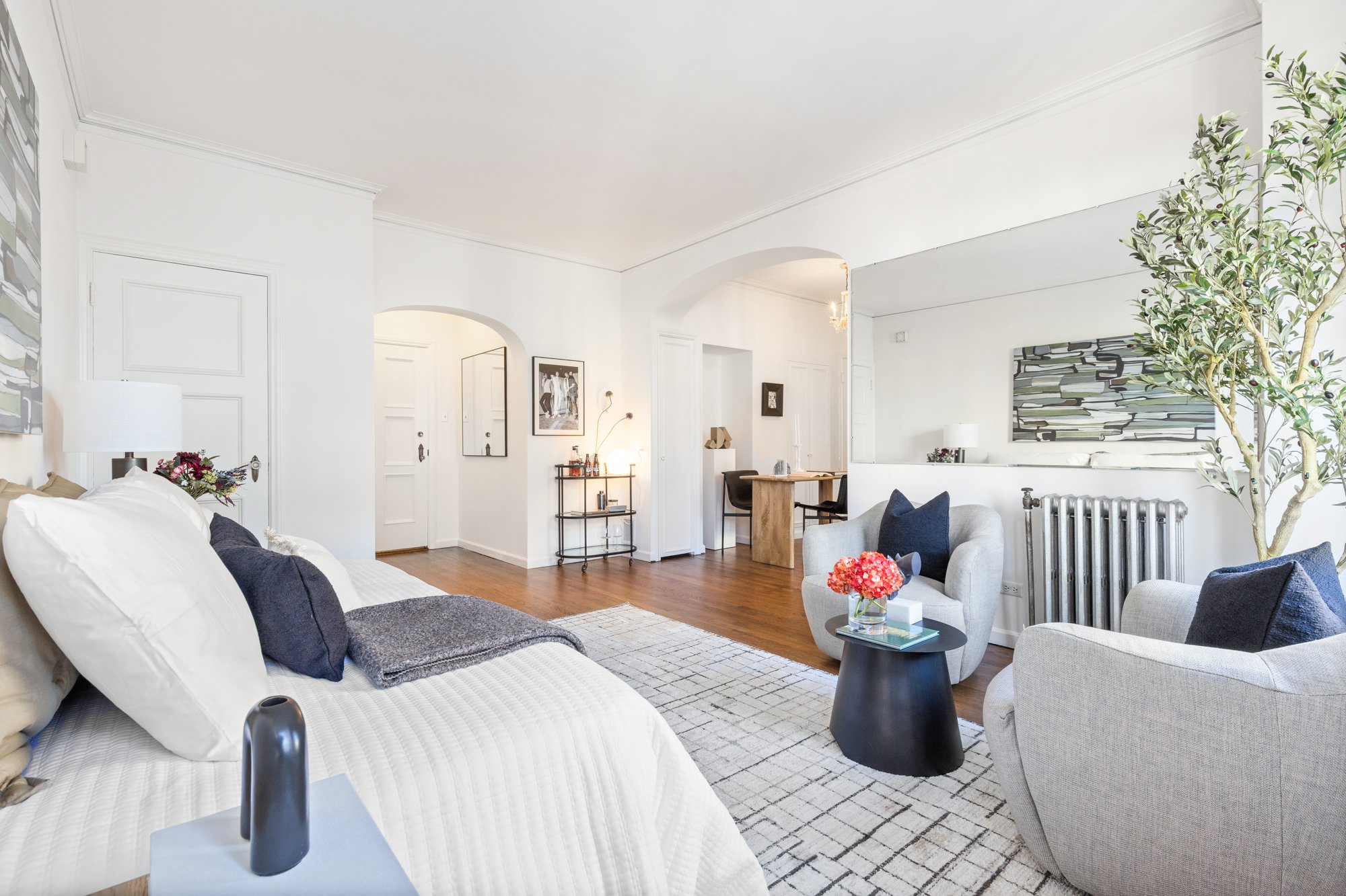 Living room with white walls, wooden floors, a white couch with dark pillows, a gray armchair, a small black side table with a glass top, large artwork on the wall, and a potted plant.