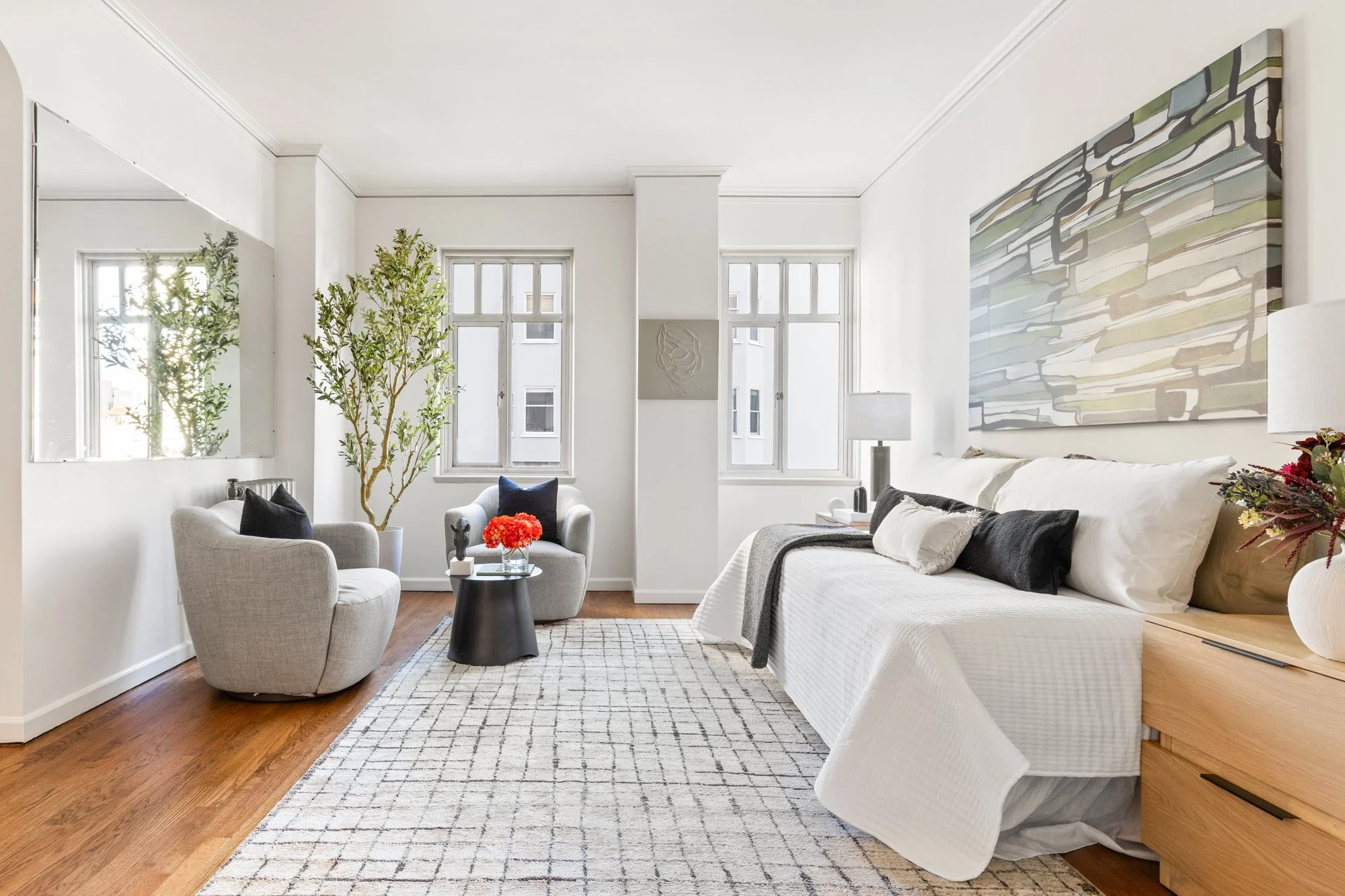Bright living room with white walls, large windows, and hardwood floors. Features include a white sofa, a small round black coffee table with a vase of red flowers, two armchairs, a large abstract painting, and a tall plant near the windows.