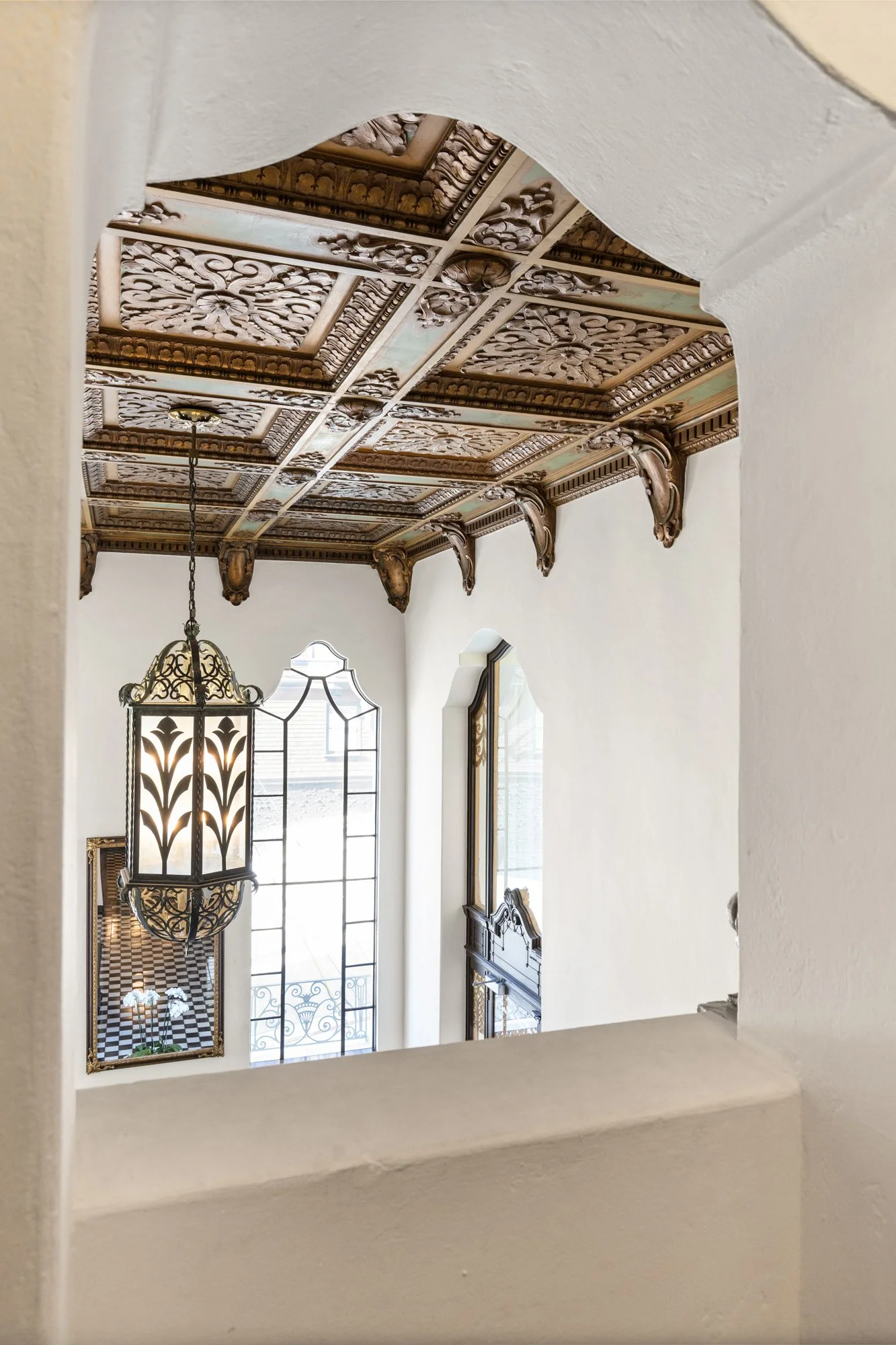 Interior view of a staircase landing with decorative ceiling, hanging lantern, arched windows with black iron fencing, and a mirror reflecting a checkered floor and potted plant.