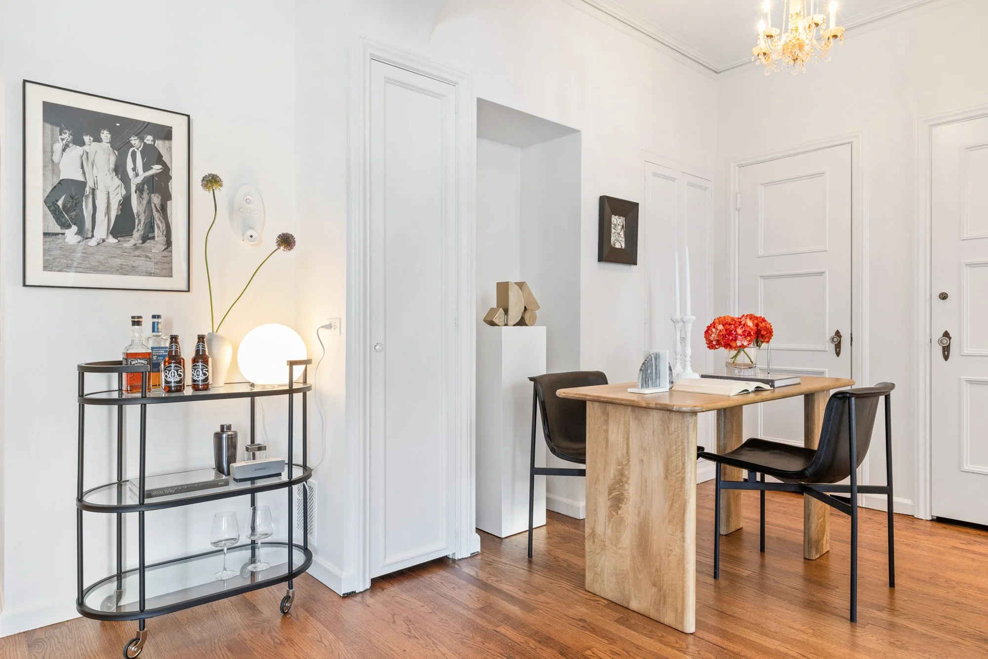 Interior of a dining area with a wooden table, black chairs, and decor including a vase with pink flowers, a small black shelf with bottles, glasses, and a lamp, and wall art with black-and-white photos. The room has white walls, wooden flooring, and