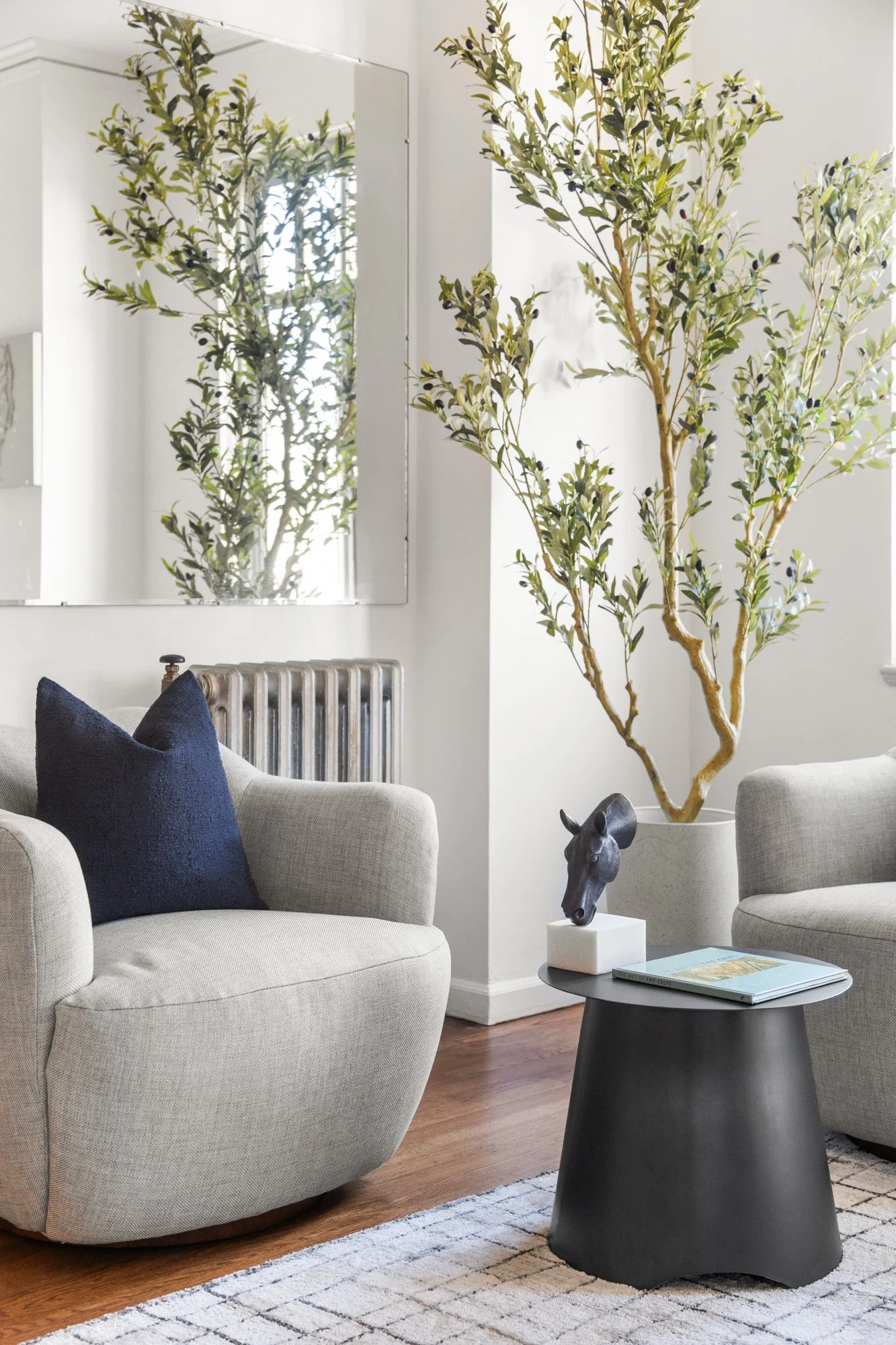 Living room with two beige armchairs, one with a navy blue pillow, a round black coffee table with a horse sculpture and books, a tall potted tree, a large mirror on the wall, and a white rug on dark wooden floors.