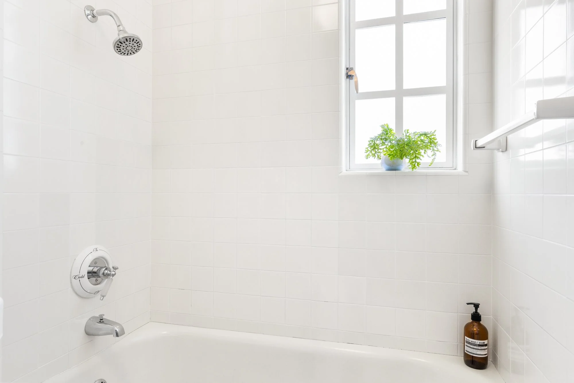 A white-tiled bathroom with a bathtub, a showerhead, a window with a potted plant, and a soap dispenser.