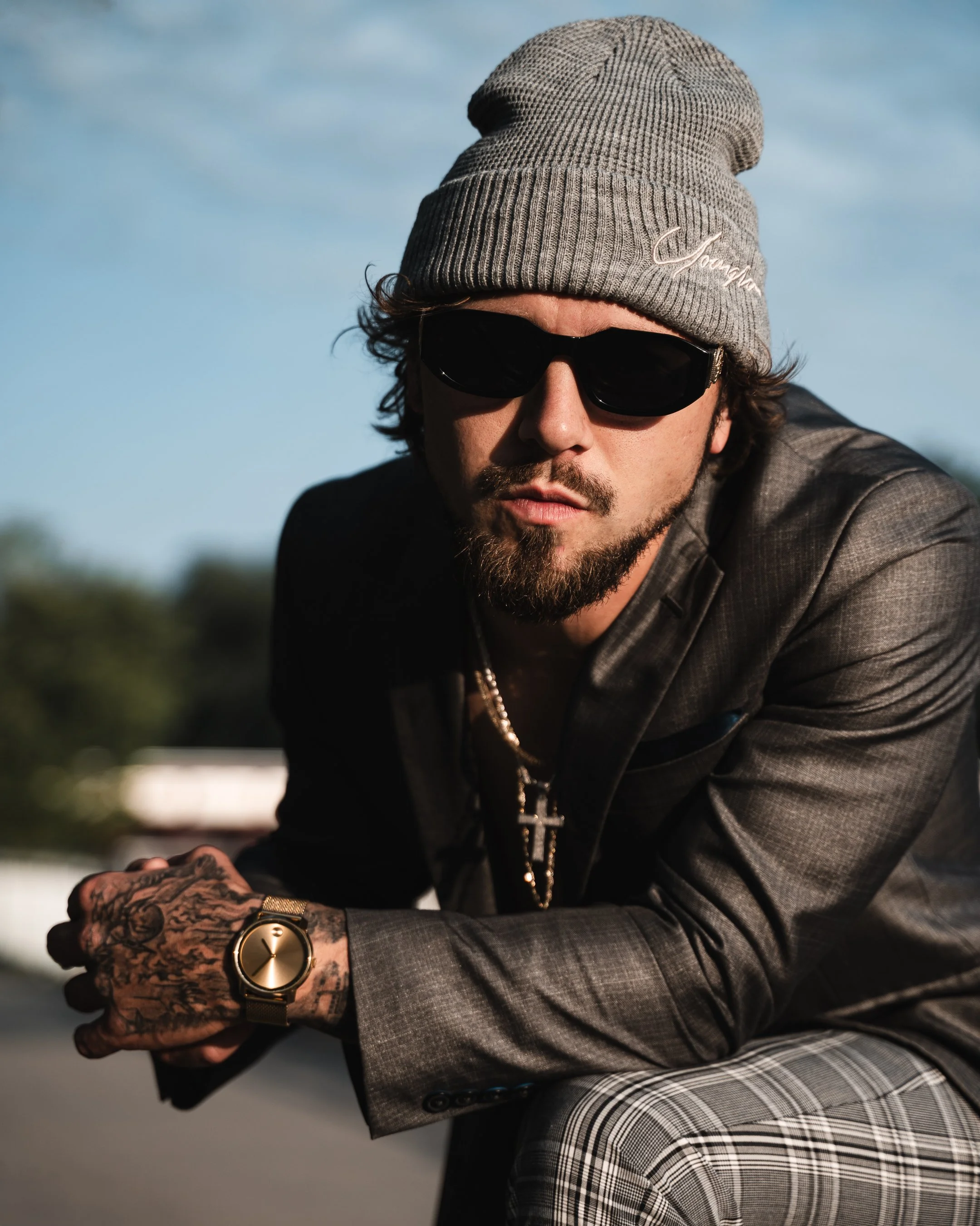 A man with dark sunglasses, a gray beanie with embroidered text, and tattoos on his hand and wrist, dressed in a gray checked blazer and gray pants, outdoors with a blurred background of clouds and greenery.