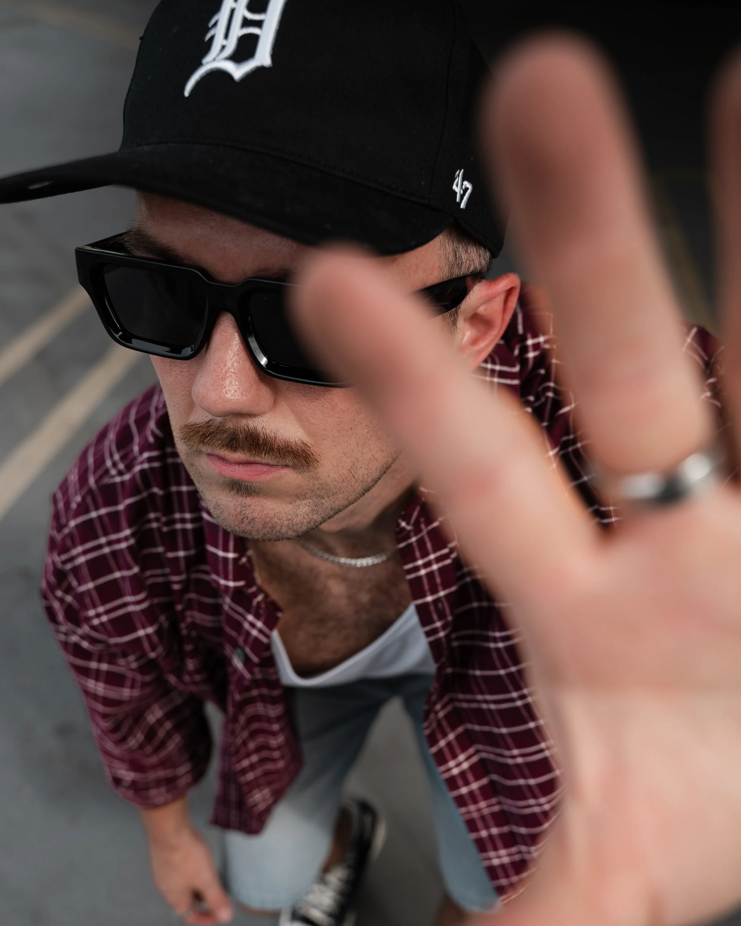 Close-up of a young man wearing a black cap with a white letter 'D', black sunglasses, a maroon plaid shirt, and a white t-shirt, taking a selfie with a slightly tilted camera, with fingers partially obscuring the view.