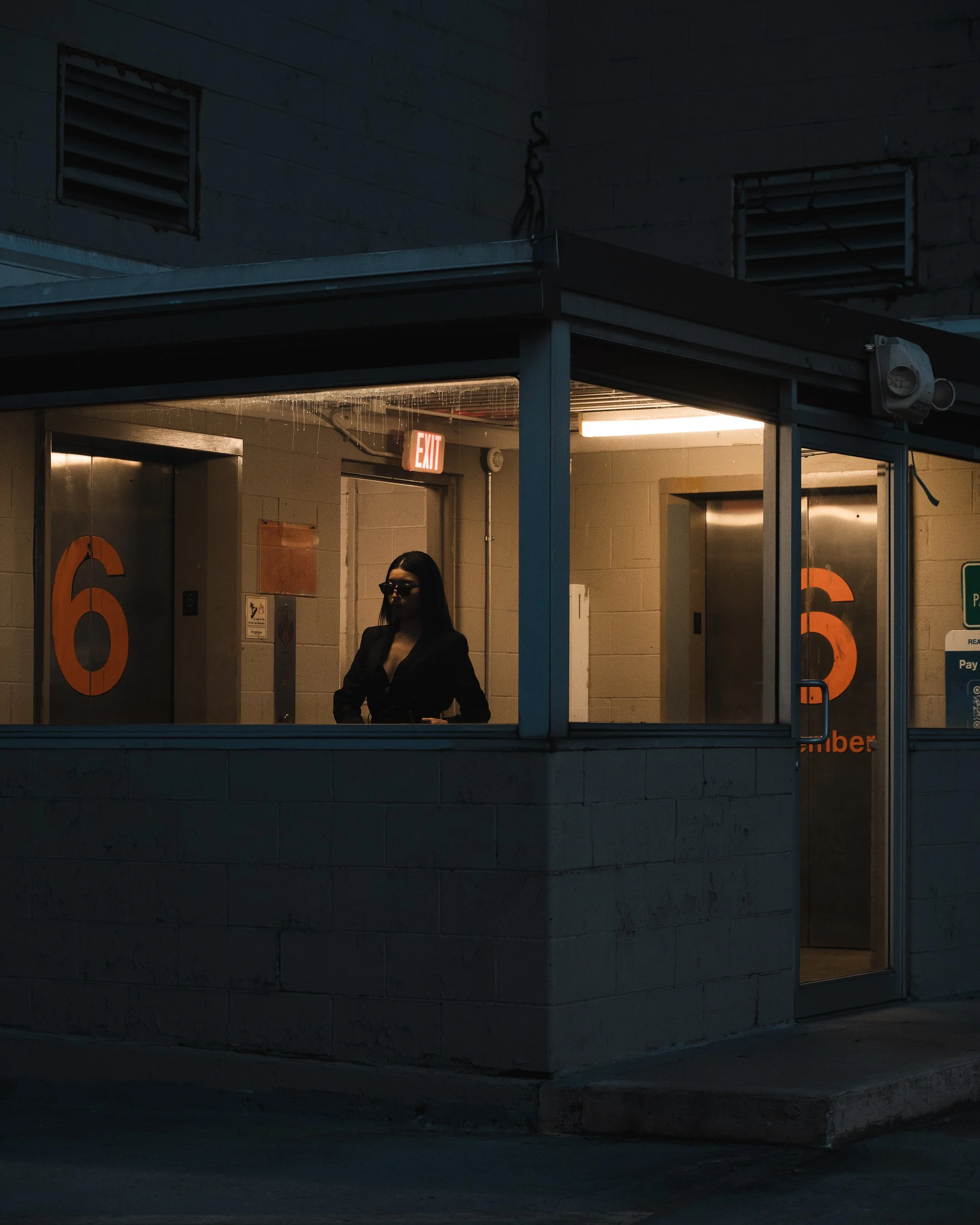 Nighttime photograph of a small building with large orange numbers 6 and 3 on the doors. A person with dark hair and glasses is standing inside near the door, with part of the person's face illuminated by interior lighting. The building has exposed b