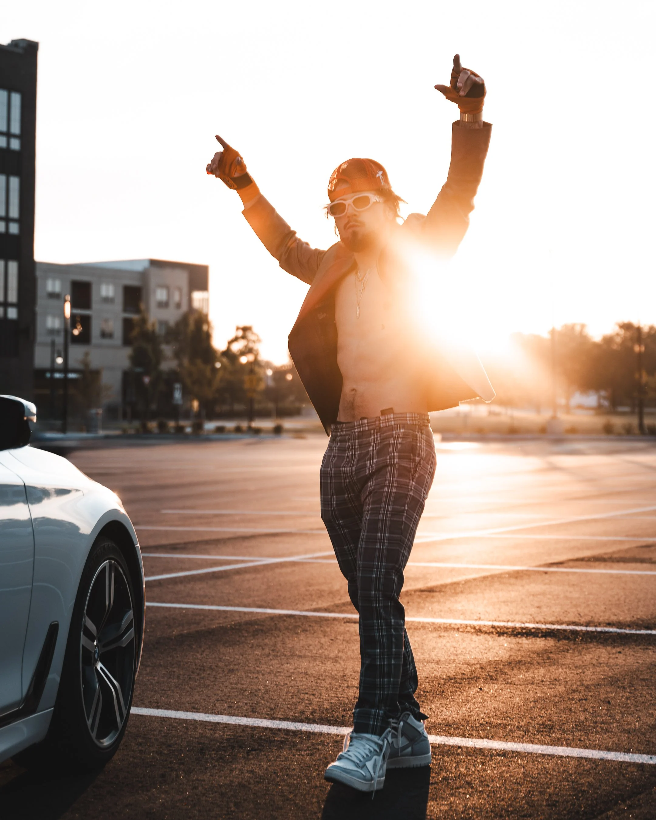 A man in plaid pants, sneakers, sunglasses, and a cap with a skateboard sticker, stands in a parking lot with his arms raised, pointing upwards during sunset with modern buildings and trees in the background.