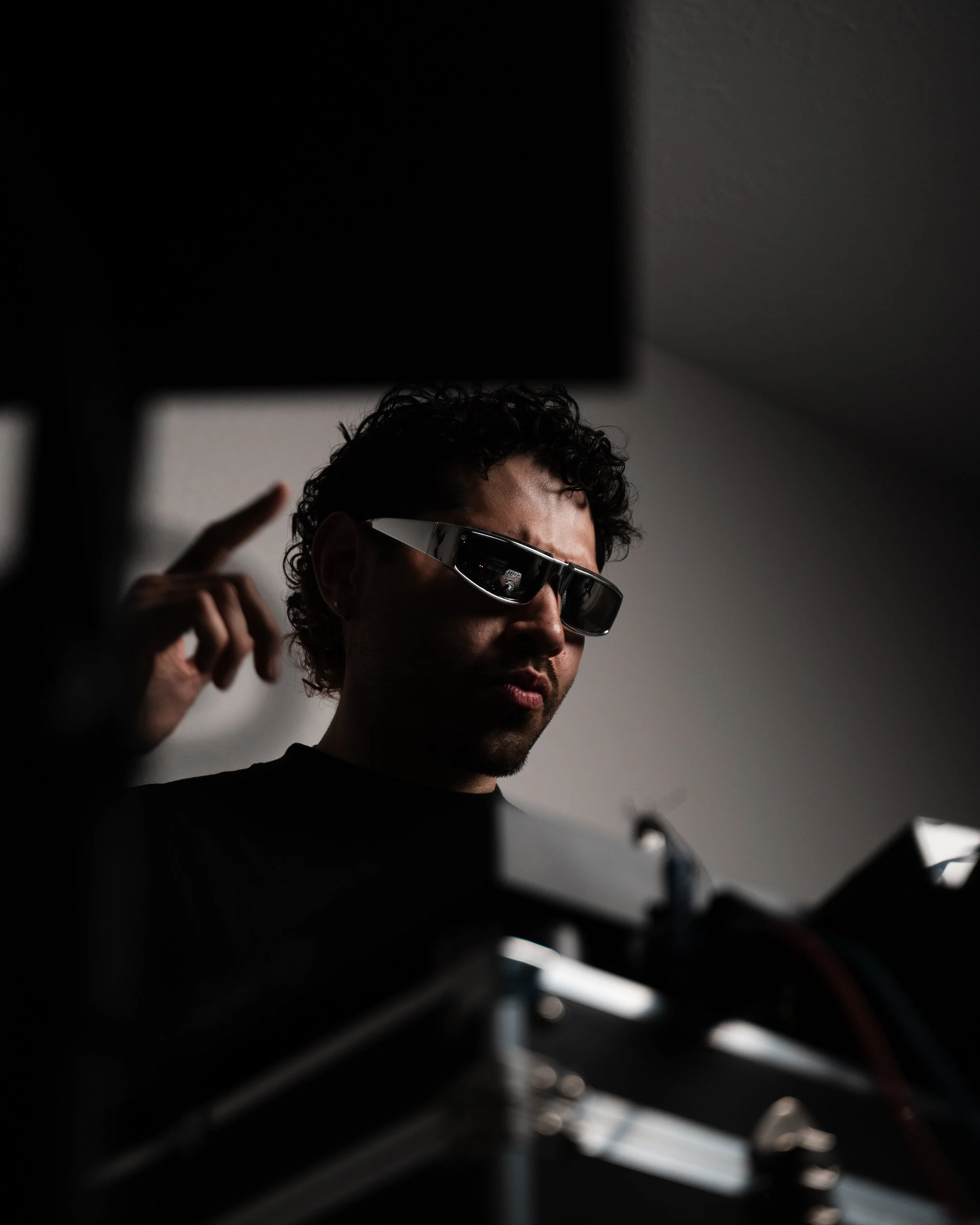 A man with curly hair wearing sunglasses, seen through a dark object with horizontal streaks, possibly a grill or louver, against a plain light-colored background.