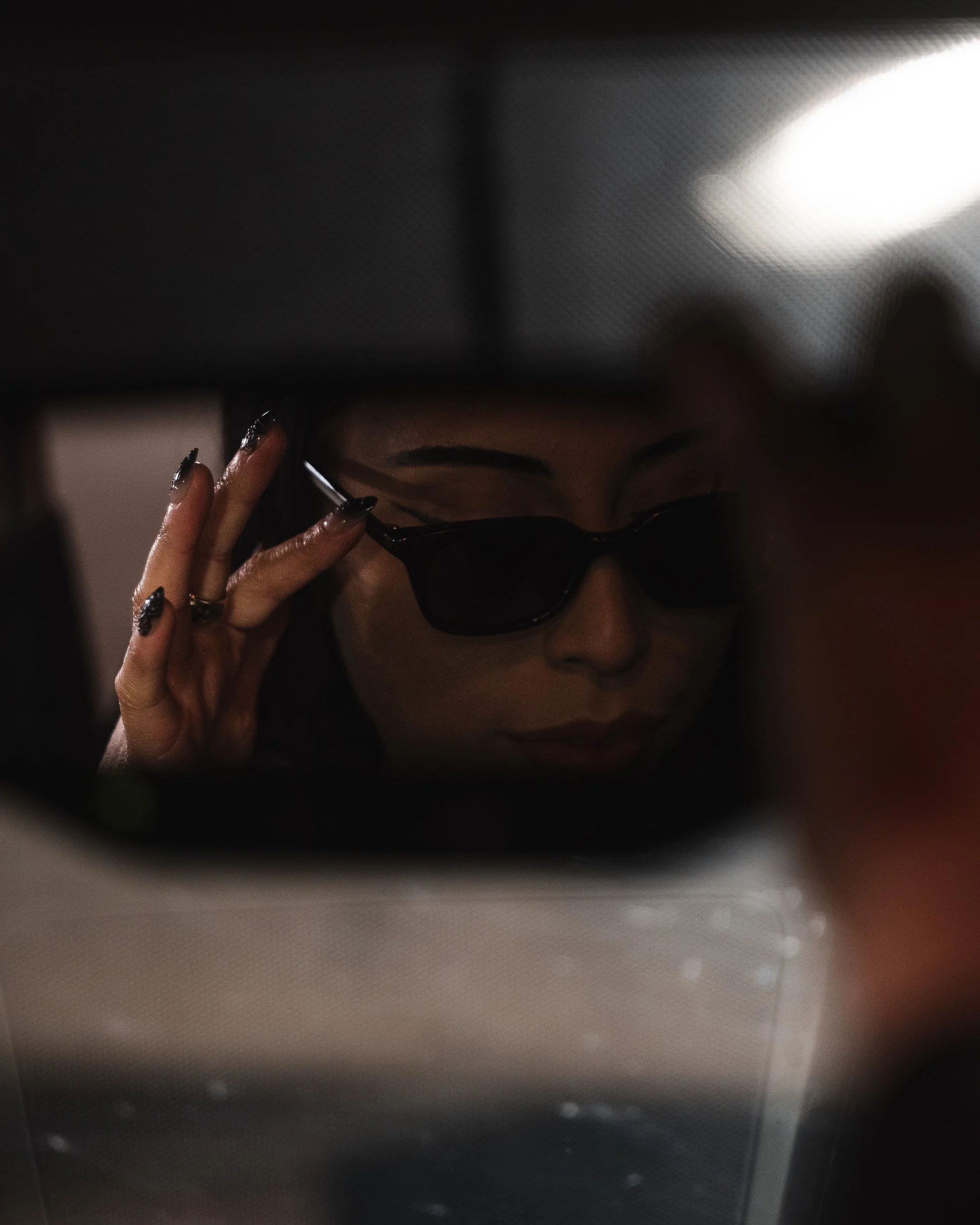 A close-up image of a person's face partially visible through a reflective, possibly metallic or glossy, surface, with the person's eyes covered by dark sunglasses and hand near the face.