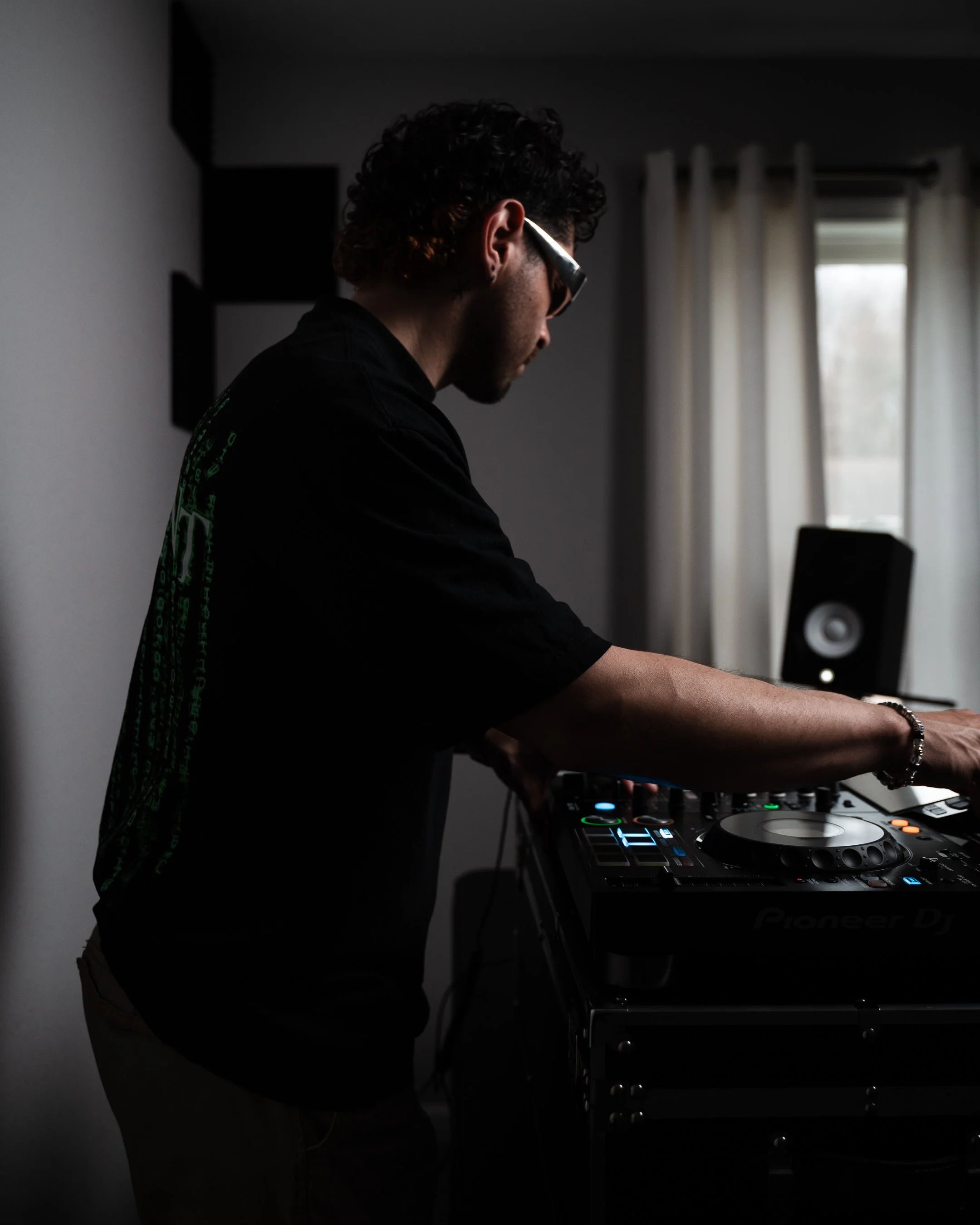 A person with curly hair and sunglasses DJing at a music mixer in a dimly lit room with a window in the background.