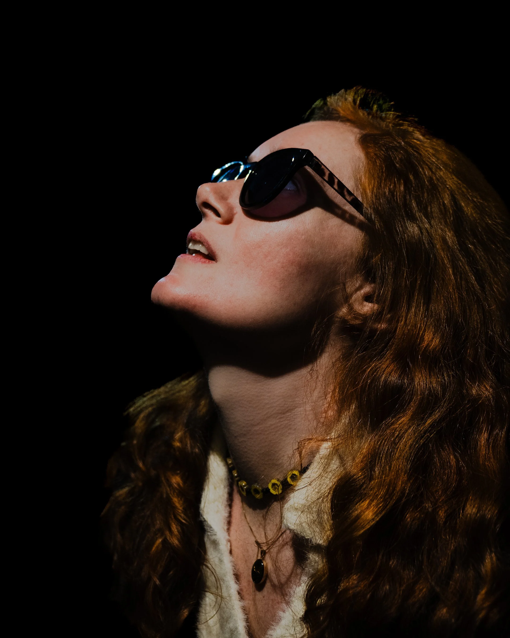 A woman with red hair wearing sunglasses, a fur coat, a beaded necklace, and a matching bracelet against a black background.