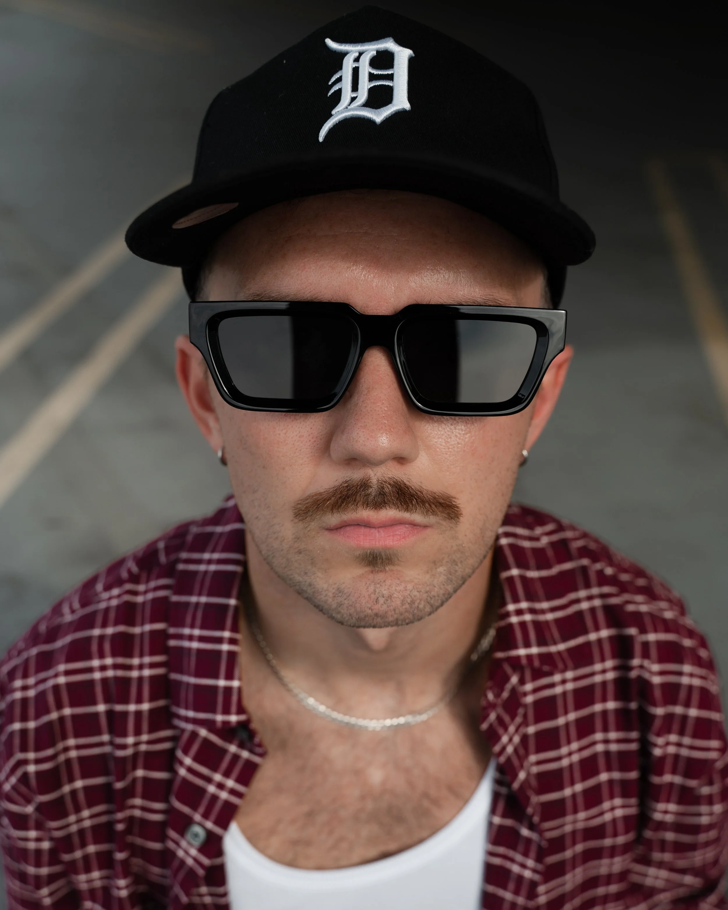 Close-up of a person wearing a black baseball cap with a white 'D' logo, black sunglasses, a red plaid shirt, and a silver chain necklace, with part of a white shirt visible underneath.