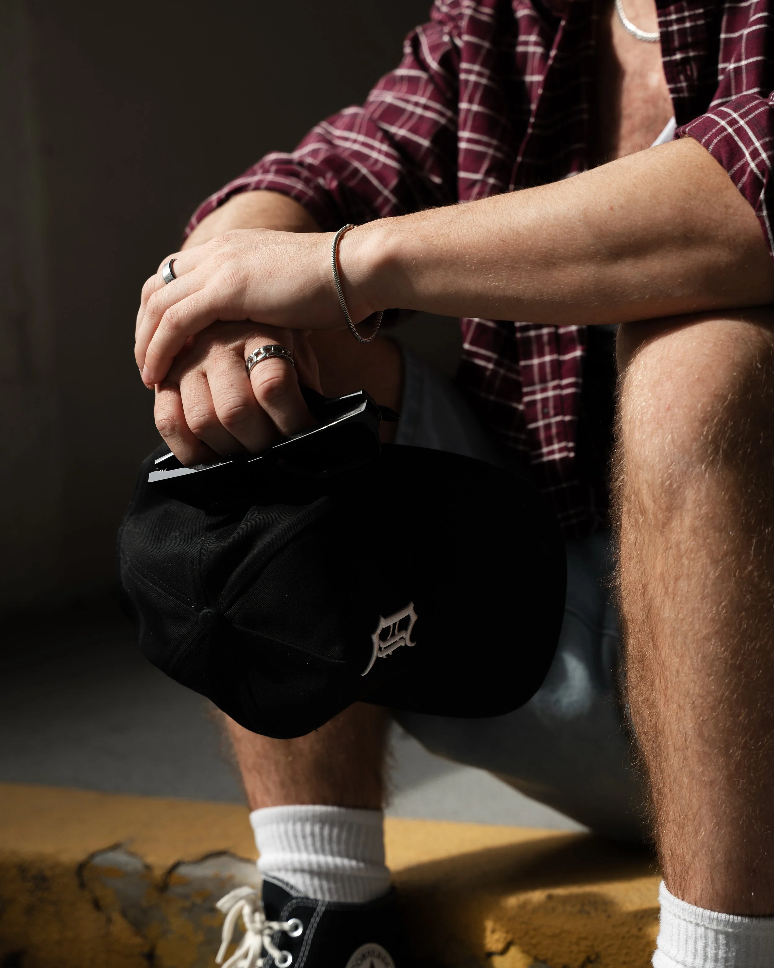 A person wearing a red plaid shirt, black shorts with a logo, white socks, and black sneakers with white laces, is squatting or kneeling on a wooden floor. Only part of the person's torso and legs are visible.