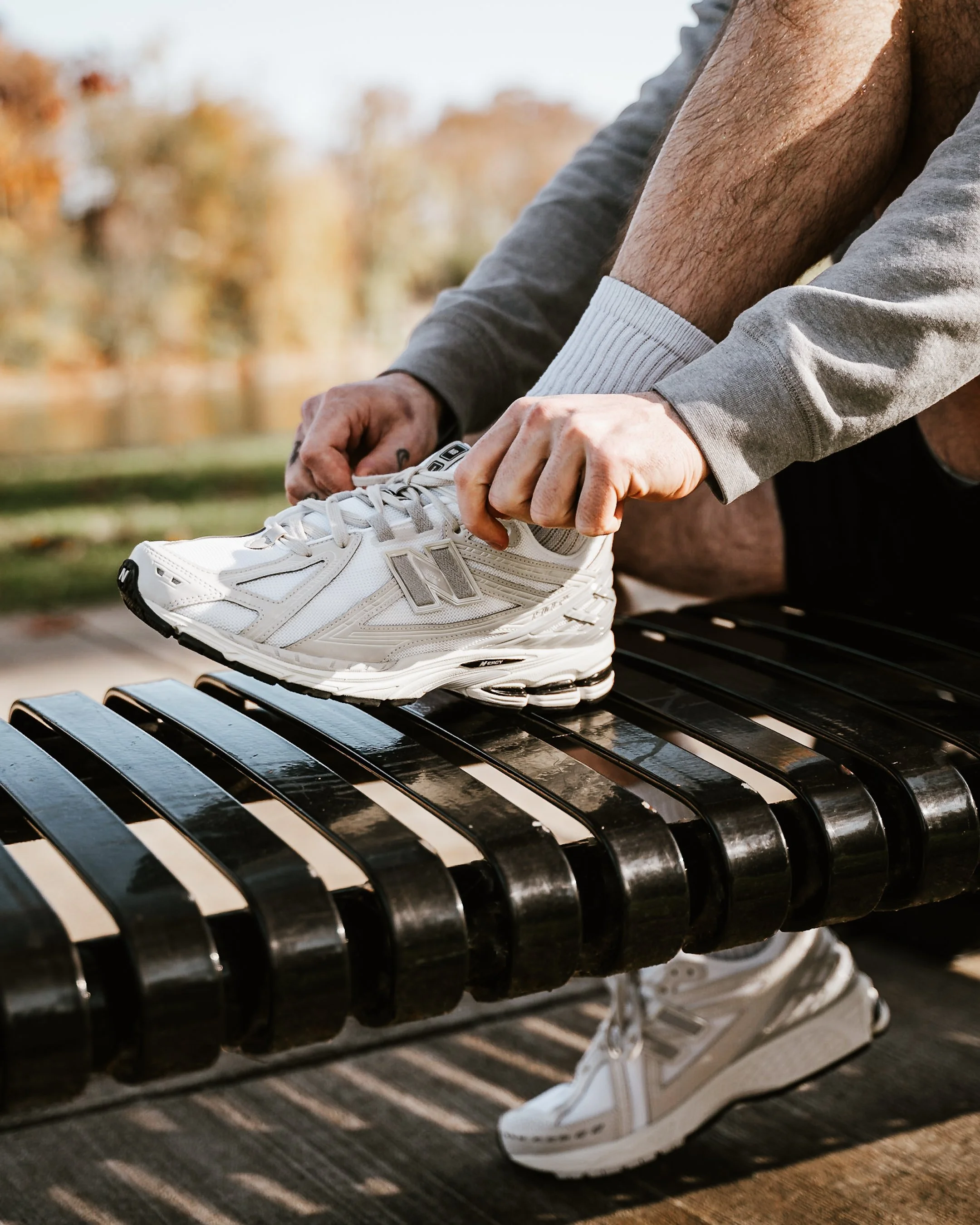 A person tying their white athletic shoe on a black metal bench outdoors with autumn trees in the background.