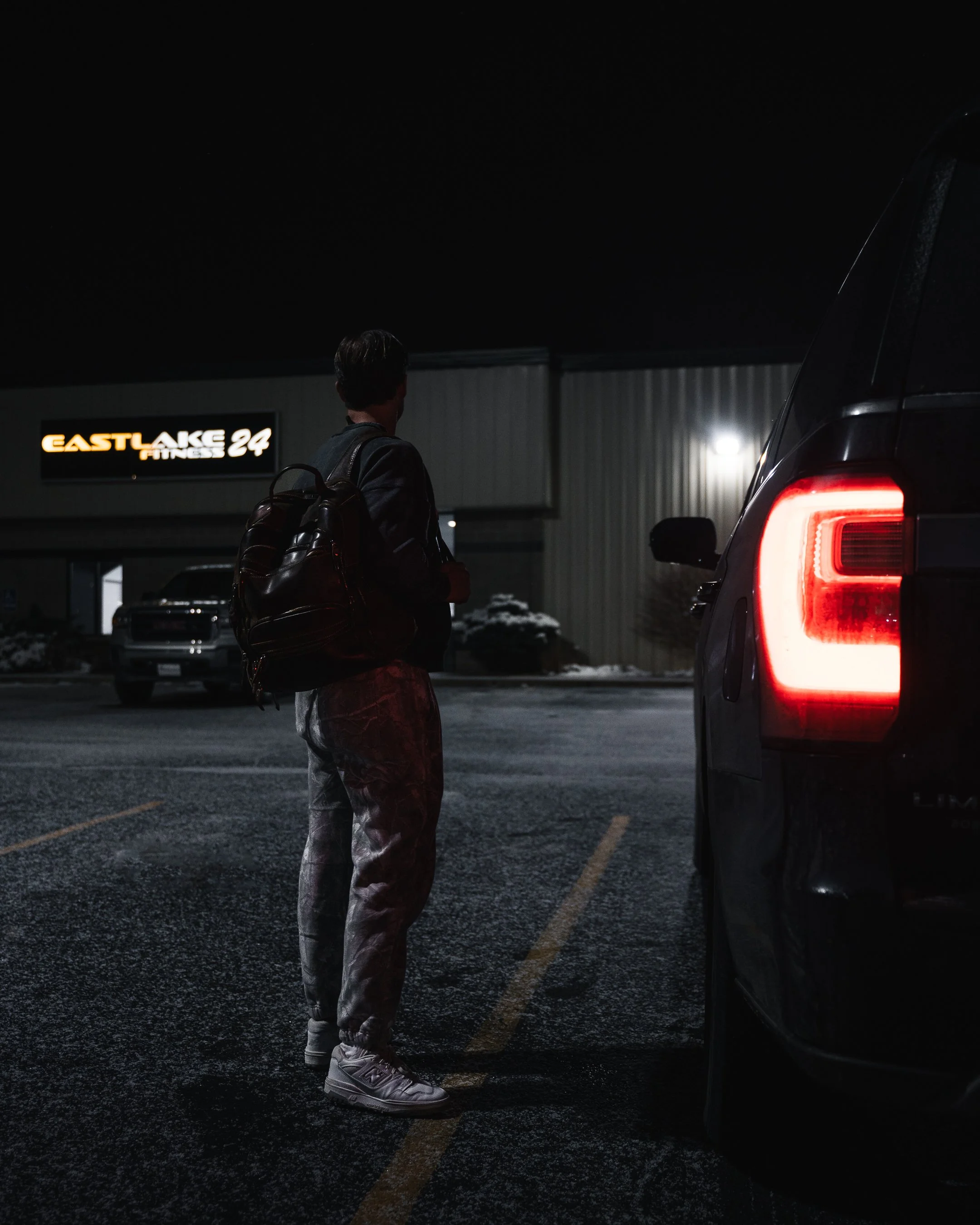 A person standing in a parking lot at night next to a large vehicle, with their back to the camera, wearing a backpack, a dark jacket, light-colored pants, and sneakers. In the background, there is a building with a sign that reads 'EASTLAKE FITNESS 