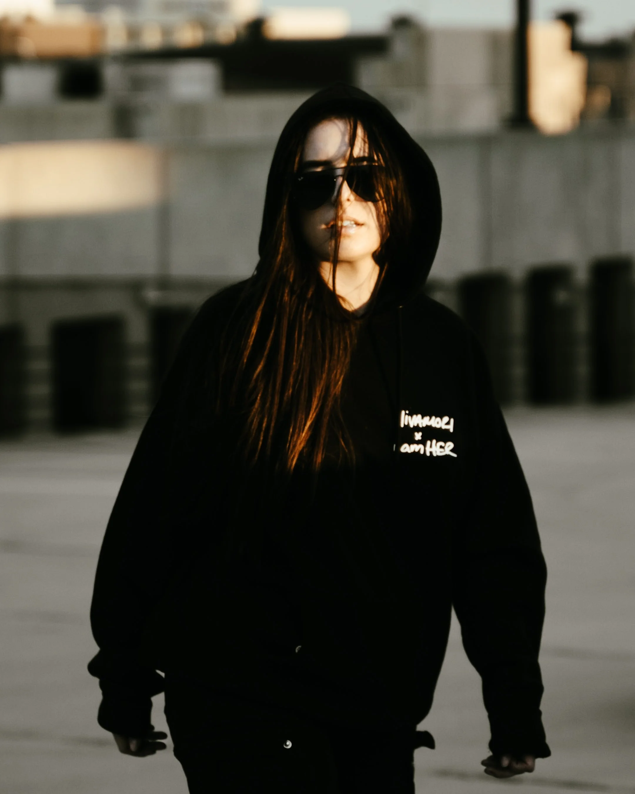 A woman with long hair and sunglasses wearing a black hoodie that says "I am HER" stands outdoors during sunset or sunrise, with blurred buildings or cityscape in the background.