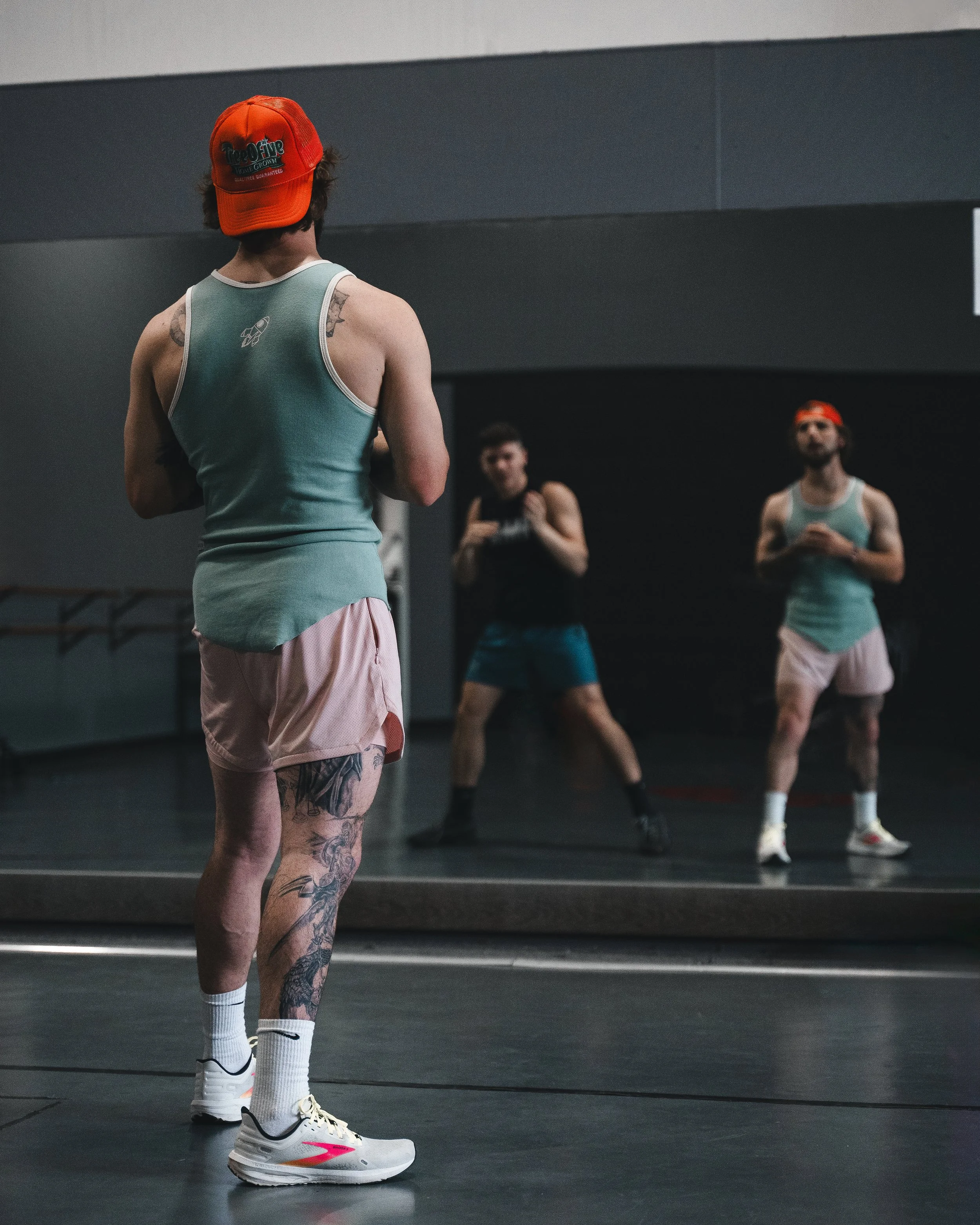 A man with tattoos on his legs and arms wearing a teal tank top, pink shorts, white sneakers with pink and yellow accents, white socks, and an orange cap, standing in front of a mirror in a gym or dance studio, facing away from the camera. In the bac