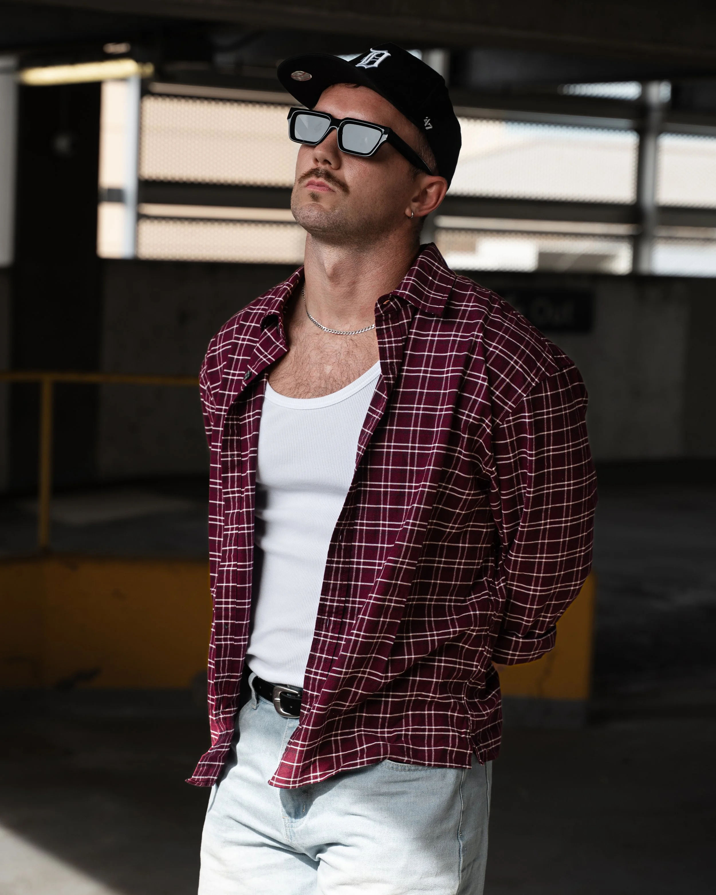 A young man with a mustache wearing a black baseball cap, black sunglasses, a silver chain, a white t-shirt, and a red plaid shirt unbuttoned, standing in a parking garage with a concrete ceiling and metal railings.