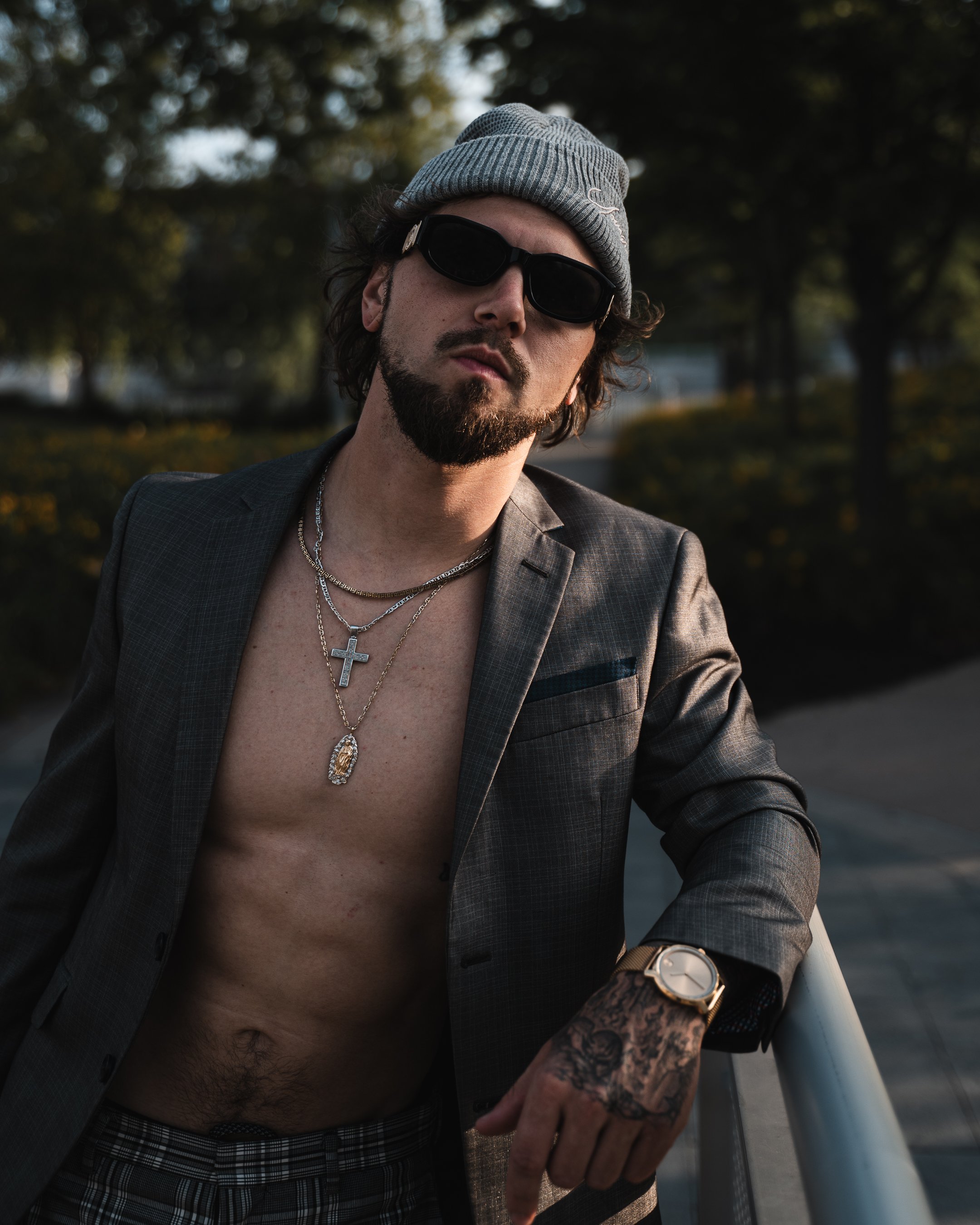 A man with a beard, wearing a gray beanie, black sunglasses, a gray checkered suit jacket, and multiple silver necklaces with pendants. He has tattoos on his hands and is looking slightly to the side while holding a surfboard. The background shows au