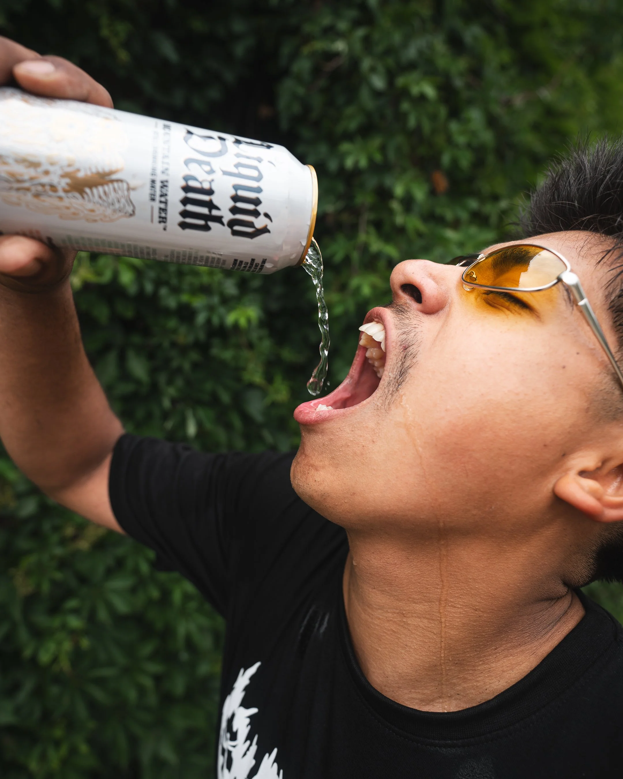 A young man with dark hair, wearing sunglasses, is pouring a cold beverage from a can into his open mouth outdoors, with green foliage in the background.
