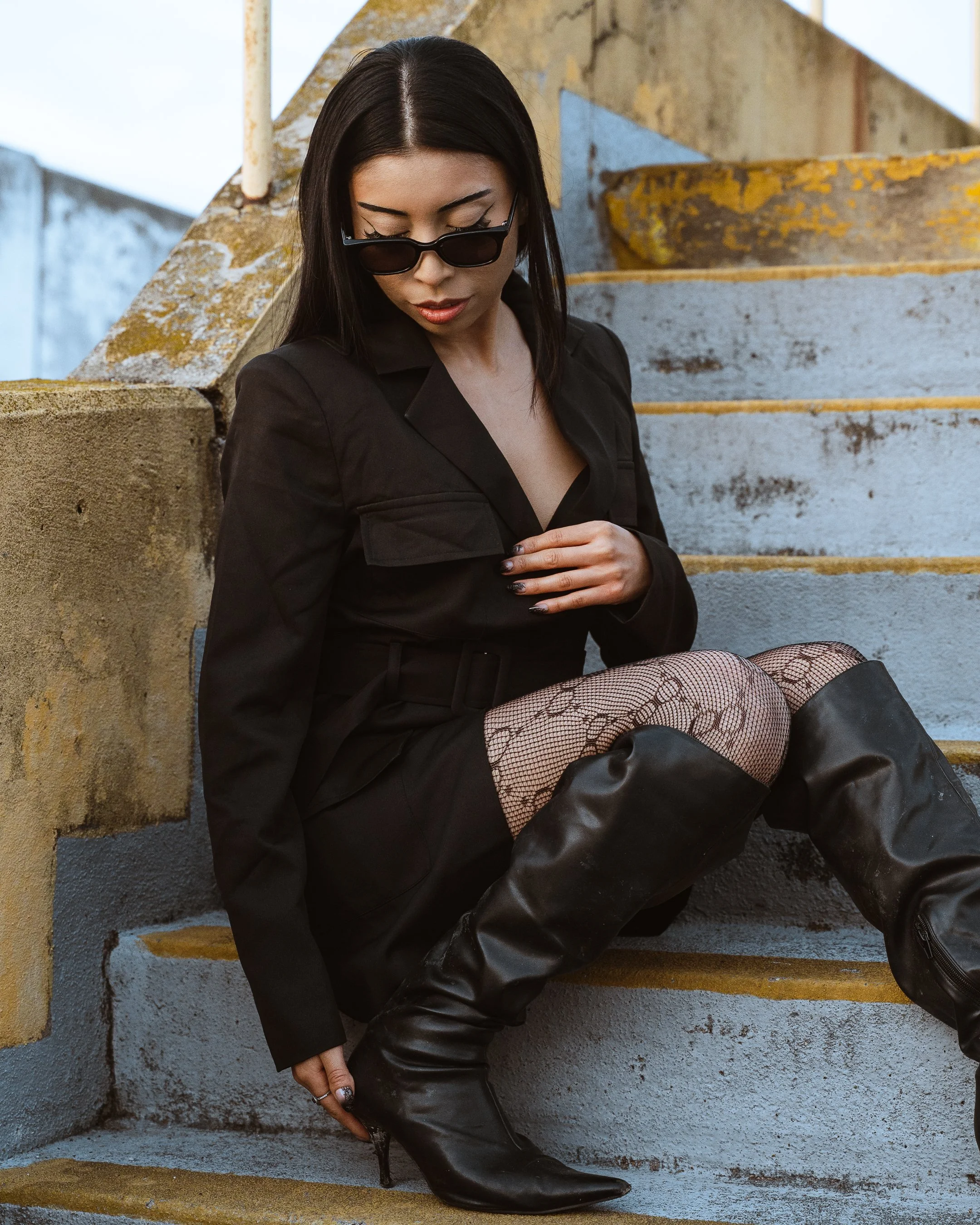 A woman sitting on concrete stairs outdoors, wearing sunglasses, a black blazer, fishnet stockings, and black over-the-knee boots.
