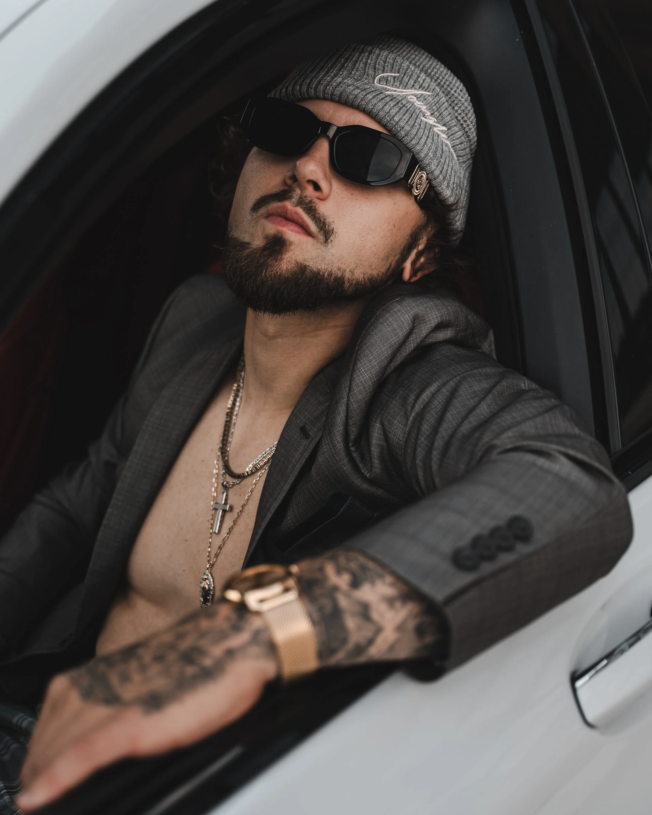 Man with a beard, sunglasses, gray beanie, and necklaces, sitting inside a vehicle, visible through the window.
