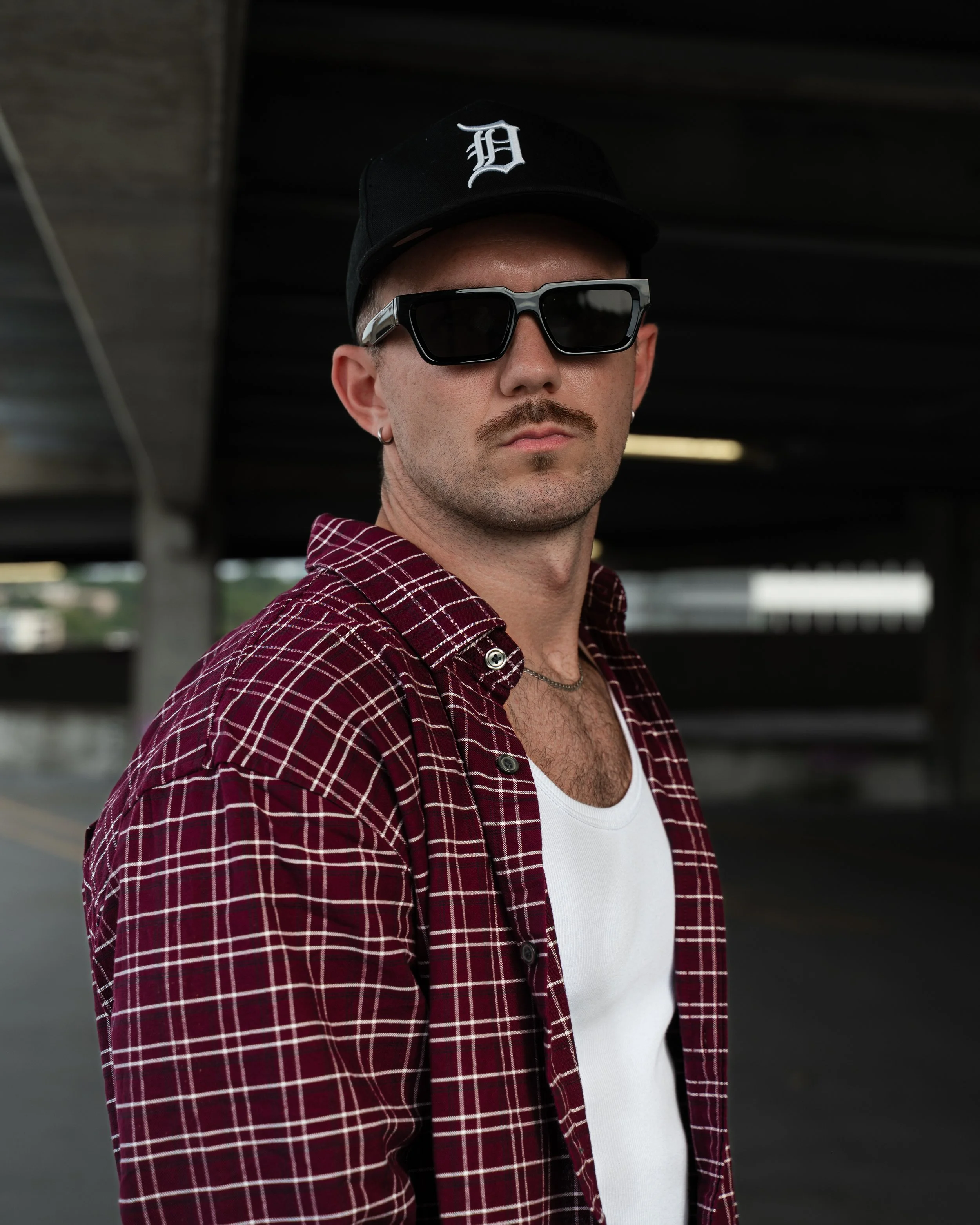 A man with light skin, wearing sunglasses, a black baseball cap with a white 'D' logo, a red plaid shirt, and a white t-shirt underneath, standing in an urban setting with dark overhead structures.