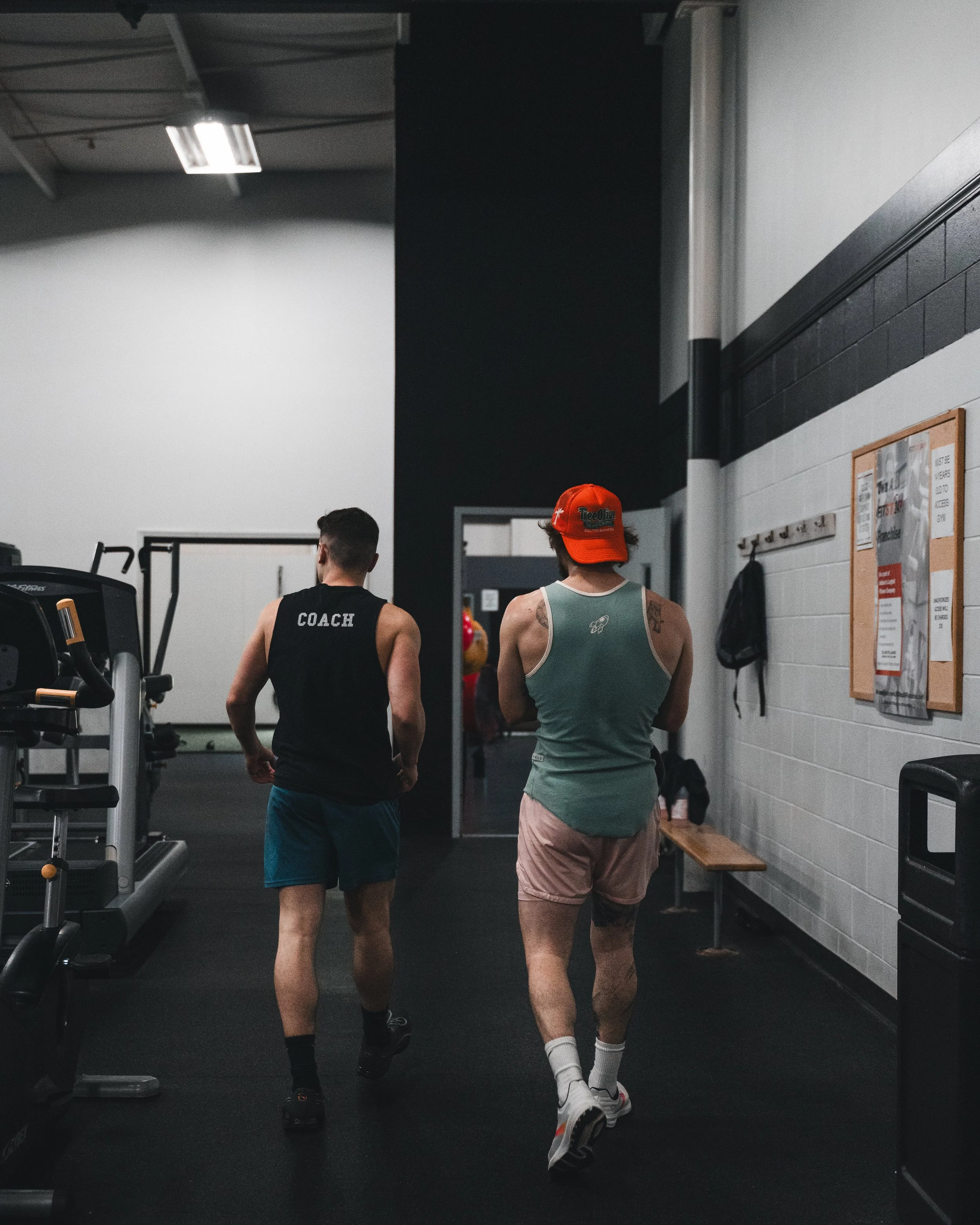 Two men with athletic builds walking inside a gym, one wearing a black sleeveless shirt with 'COACH' printed on the back and the other in a green tank top and tan shorts, with the man in the green tank top wearing a red baseball cap.