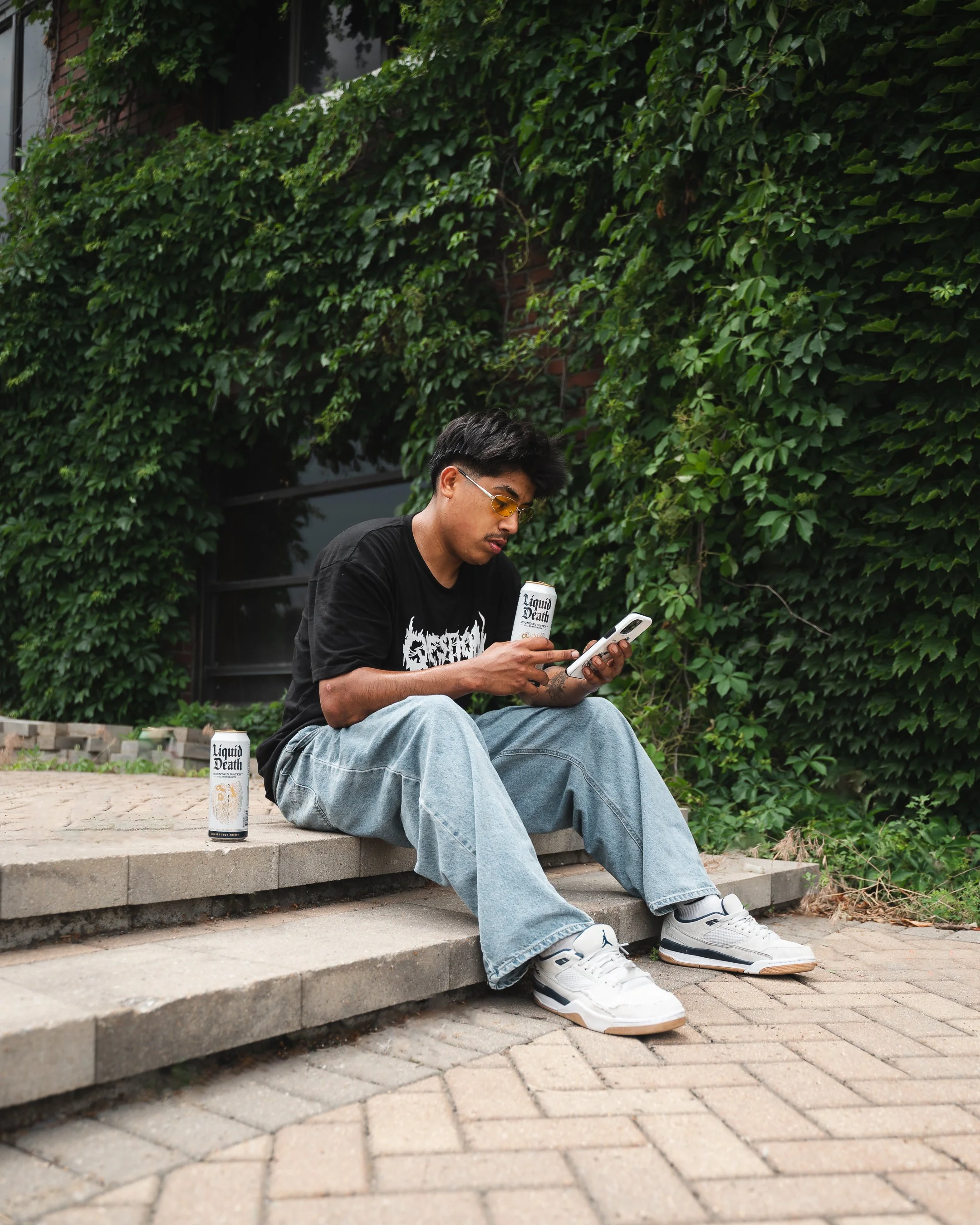 A man wearing a black shirt, light gray jeans, white sneakers, and yellow-tinted glasses is sitting on concrete steps outdoors. He is looking at his phone and holding a can of Liquid Death mountain water. There are more cans on the ground beside him,