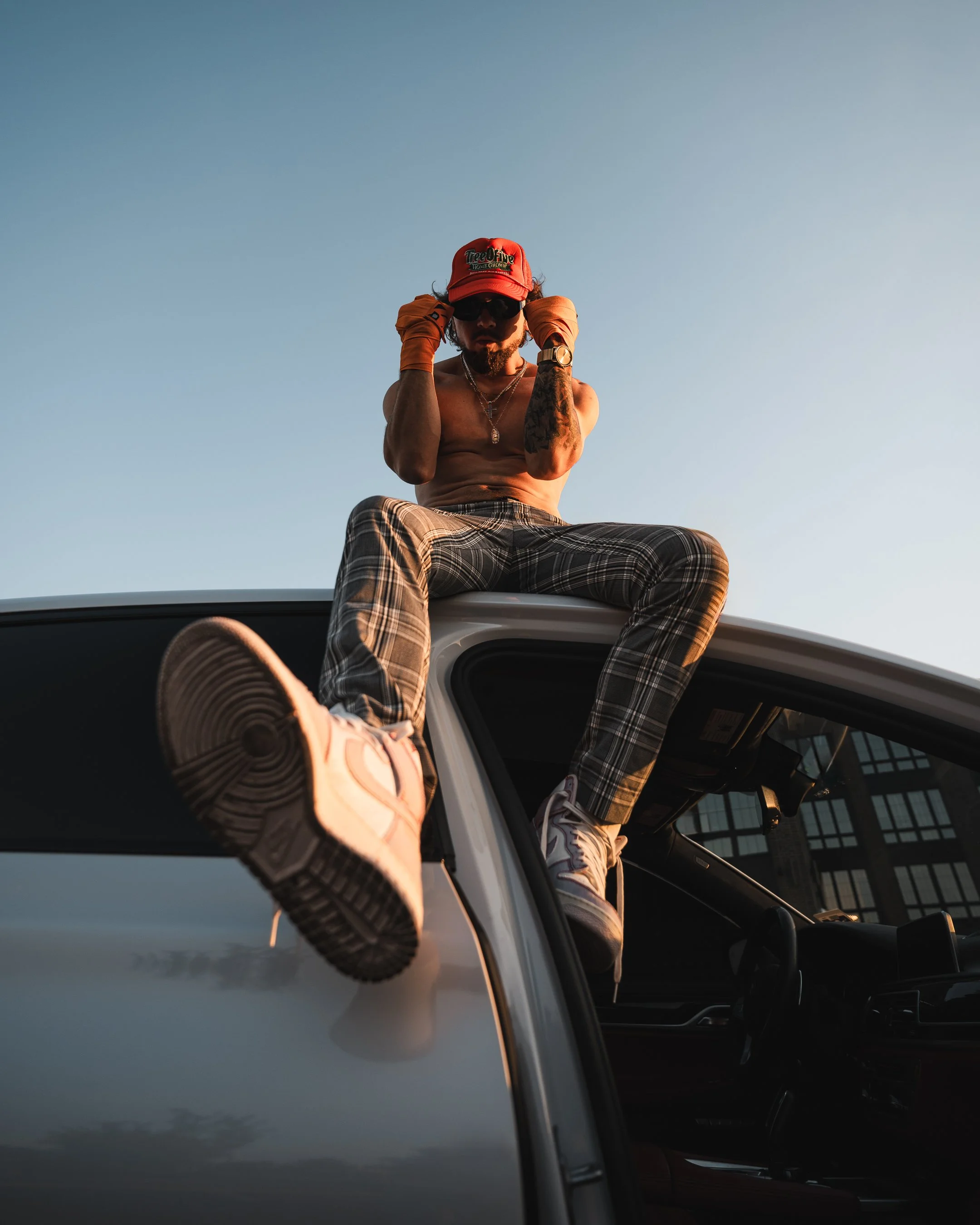 A shirtless man wearing sunglasses, a red trucker hat, and plaid pants sitting on the roof of a vehicle, adjusting his hat against a clear sky.