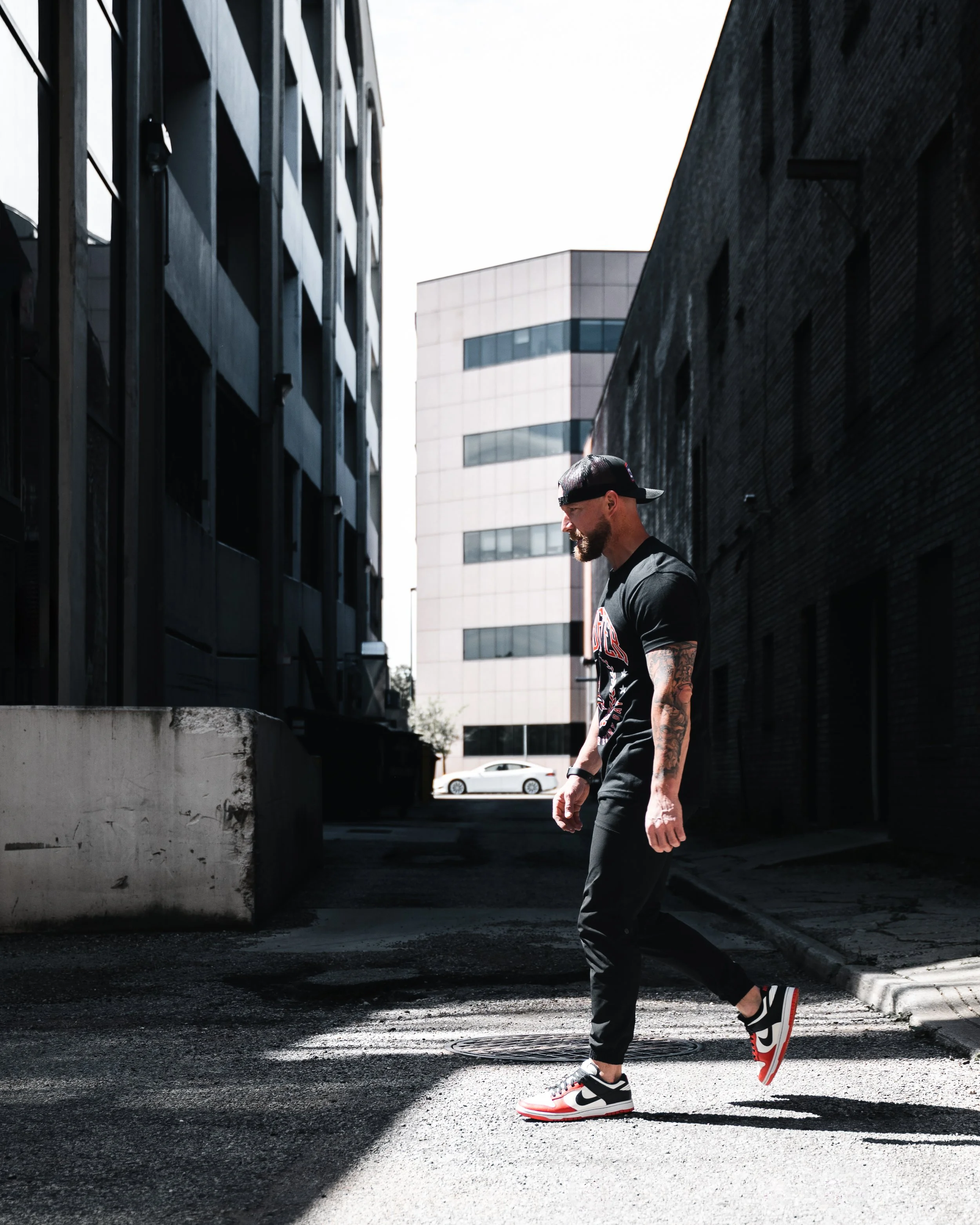 A man with tattoos on his arm, wearing a black cap backwards, black T-shirt, black pants, and red, white, and black sneakers, walking in an urban alleyway between dark buildings with another modern building in the background.