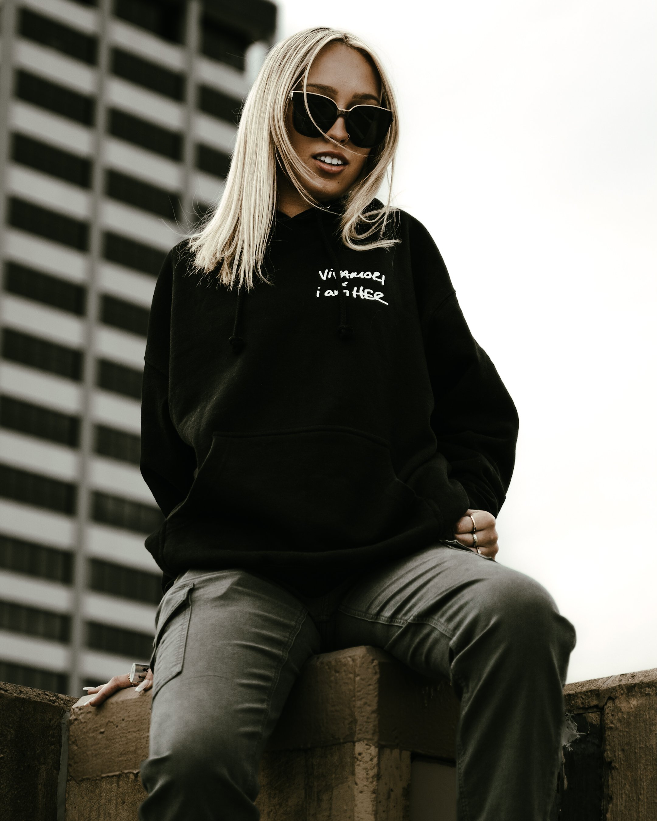 A woman with long blond hair wearing black sunglasses and a black hoodie with white text, sitting on a brick ledge outdoors in front of a tall building with a grid-like facade.