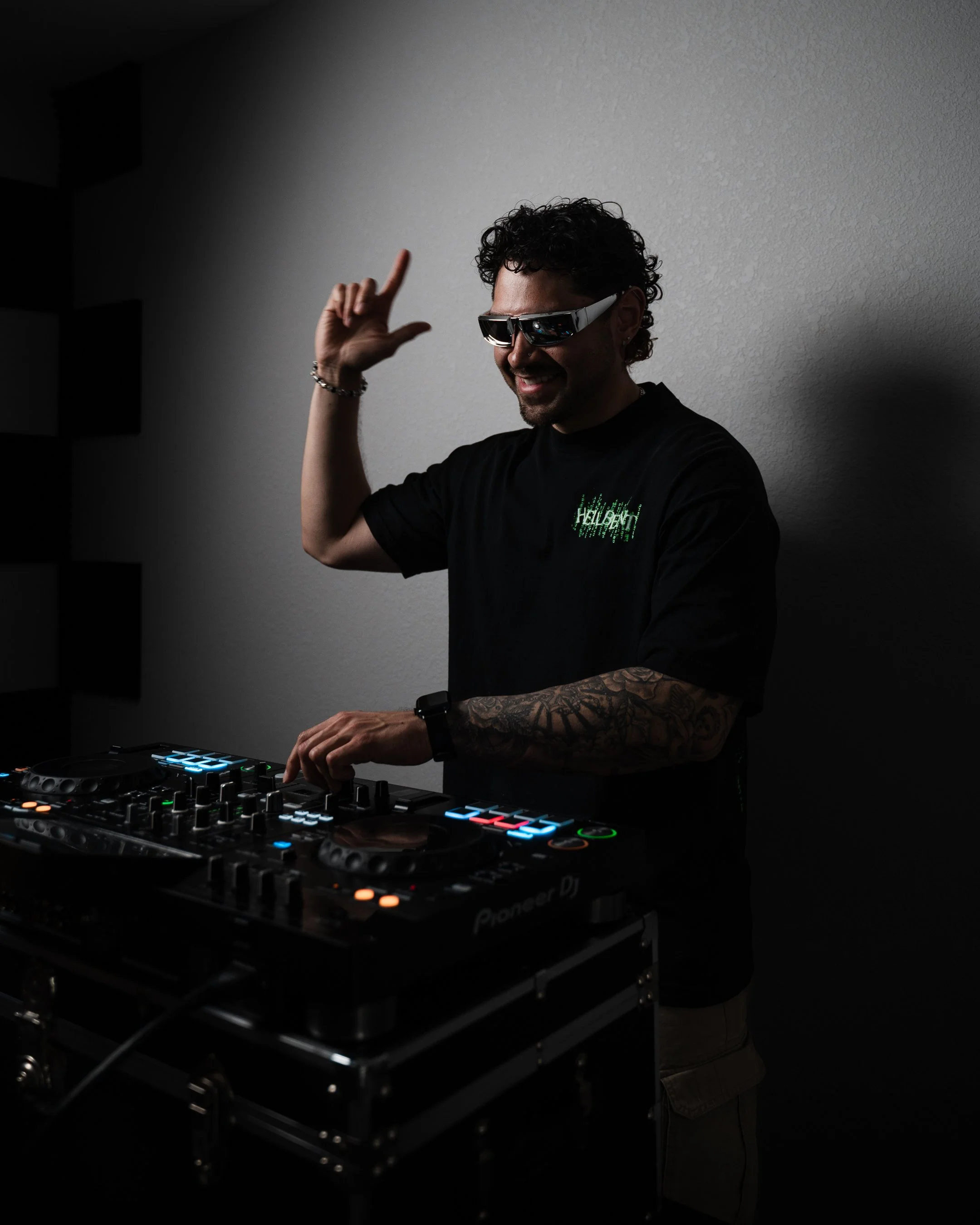 A man with curly hair, wearing sunglasses and a black t-shirt, is DJing with a Pioneer DJ controller in a dimly lit room. He is smiling and making a gesture with his right hand.