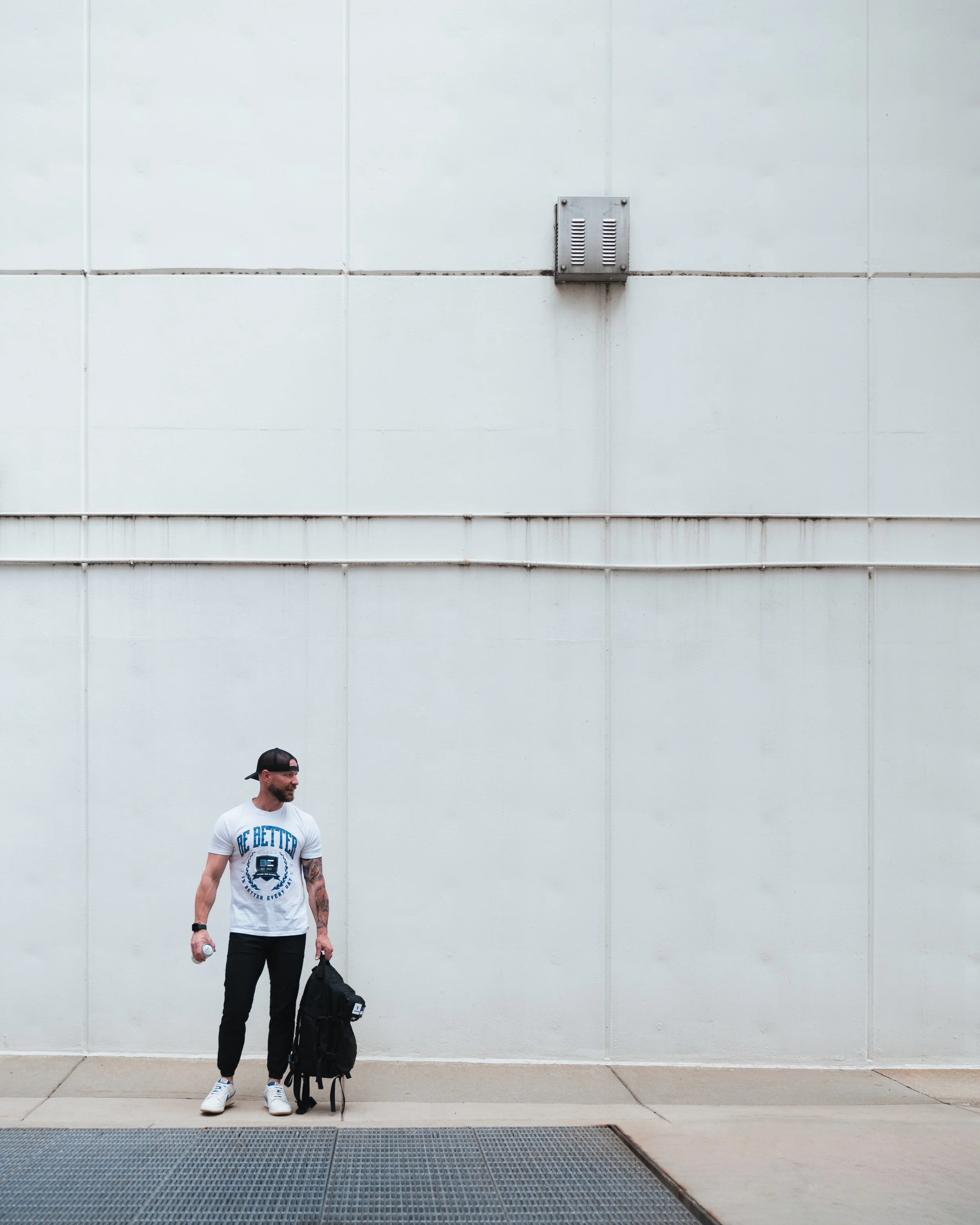 A man with a beard, wearing a white T-shirt, black pants, white sneakers, a black cap, and a watch, standing outdoors against a plain white wall. The man is holding a small bottle in his right hand and has a black backpack placed on the ground beside