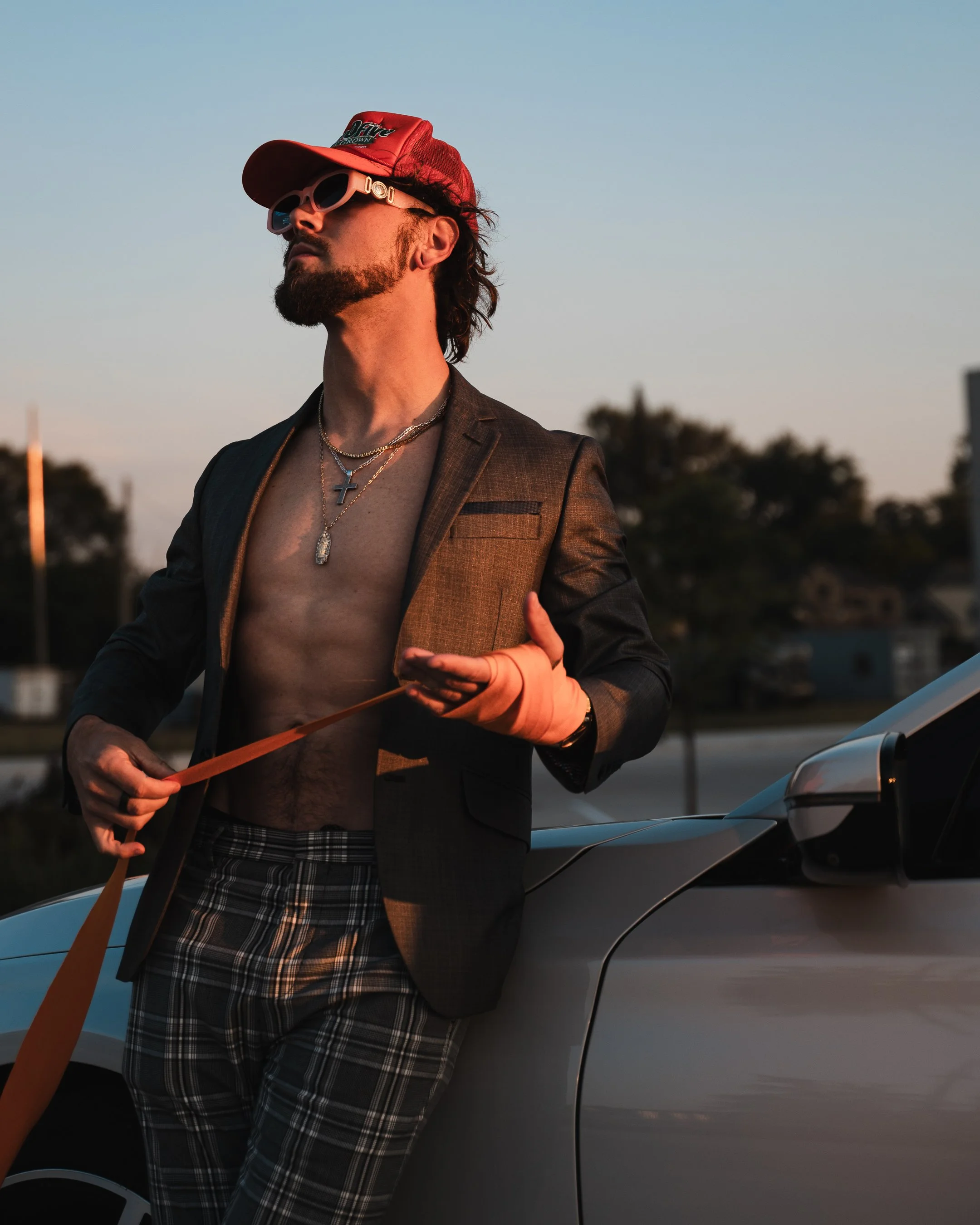 A man with a beard and long hair, wearing a red cap, pink sunglasses, a blazer, and plaid shorts, stands near a car holding a leash. The background shows a cityscape during sunset.