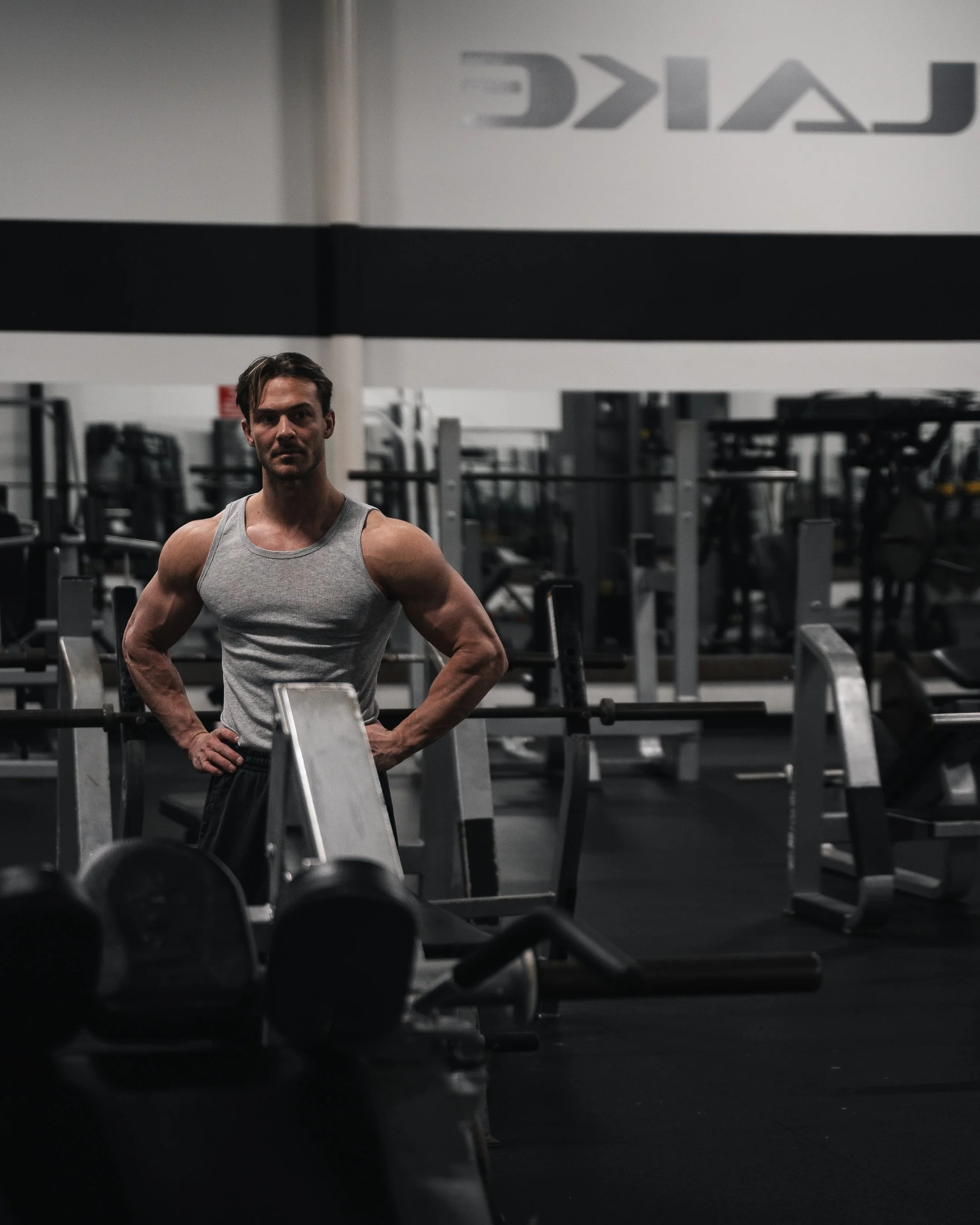 A man in a gray tank top standing in a gym surrounded by workout equipment and dumbbells, with the word 'CALIST' visible in the background.