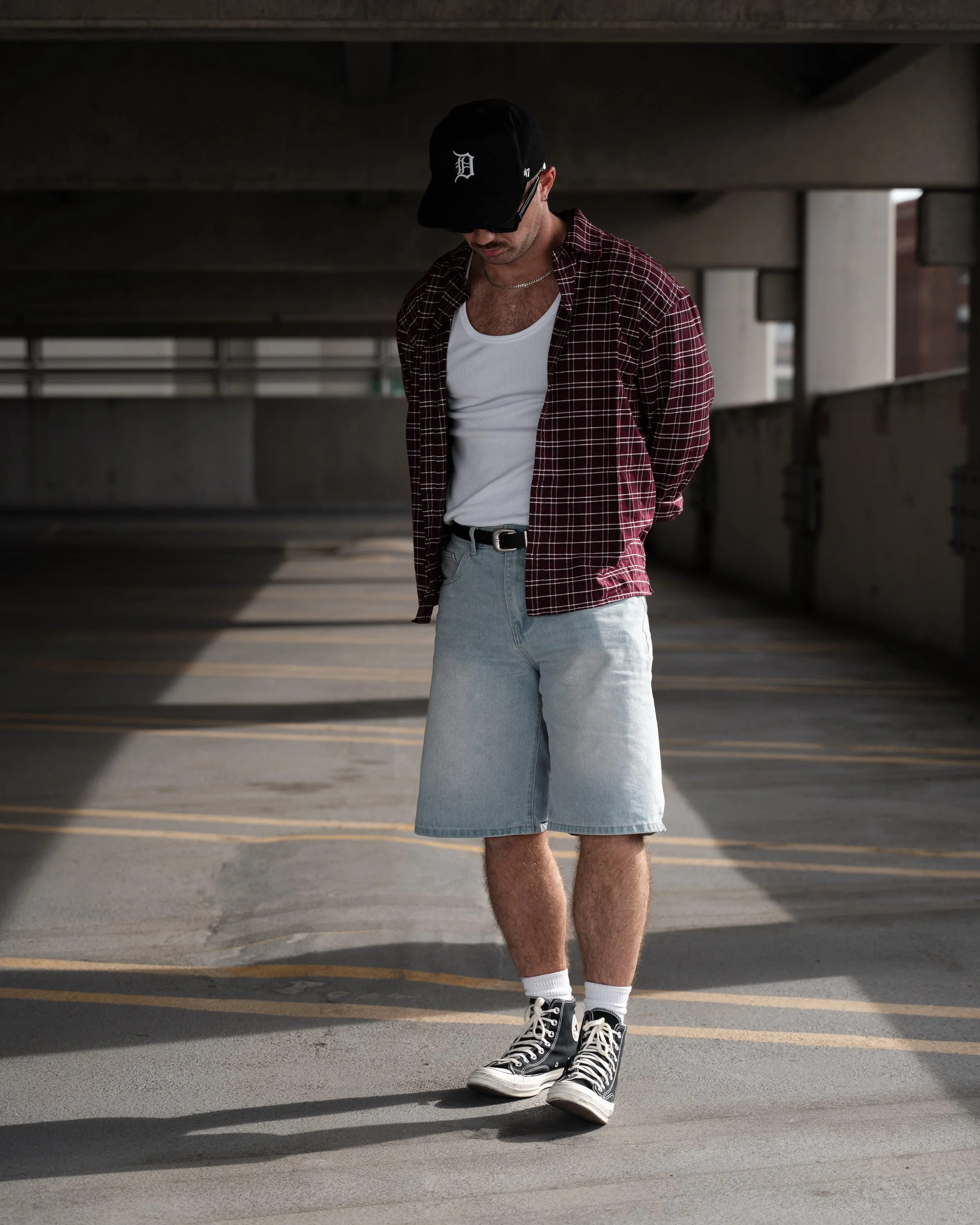 A young man wearing a black baseball cap, sunglasses, a plaid shirt over a white tank top, light denim shorts, white socks, and black high-top sneakers, standing under a concrete parking structure with sunlight casting shadows on the ground.