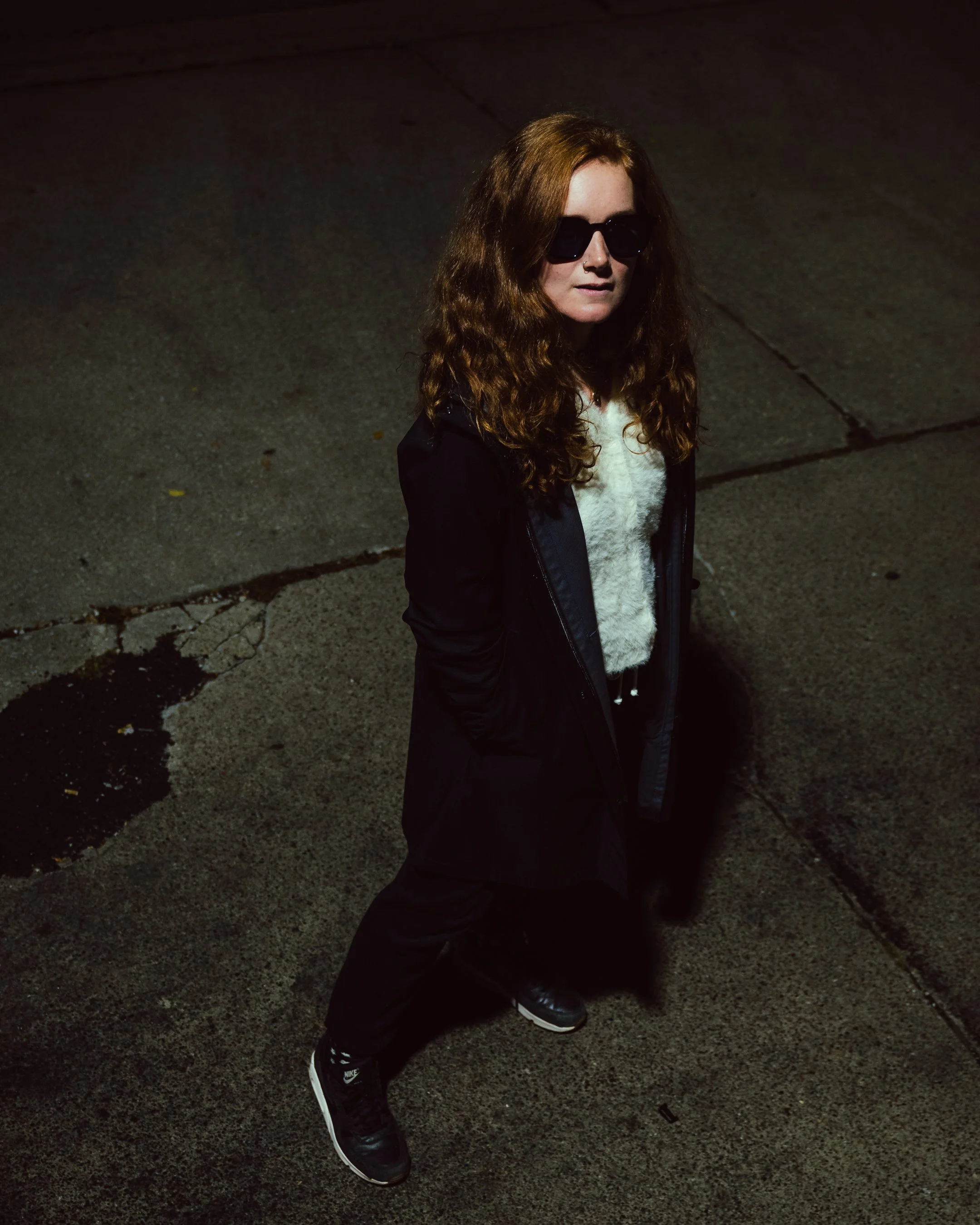 A young woman with long, curly red hair wearing dark sunglasses, a white fuzzy sweater, black jacket, black pants, and black Nike sneakers standing on a sidewalk at night.