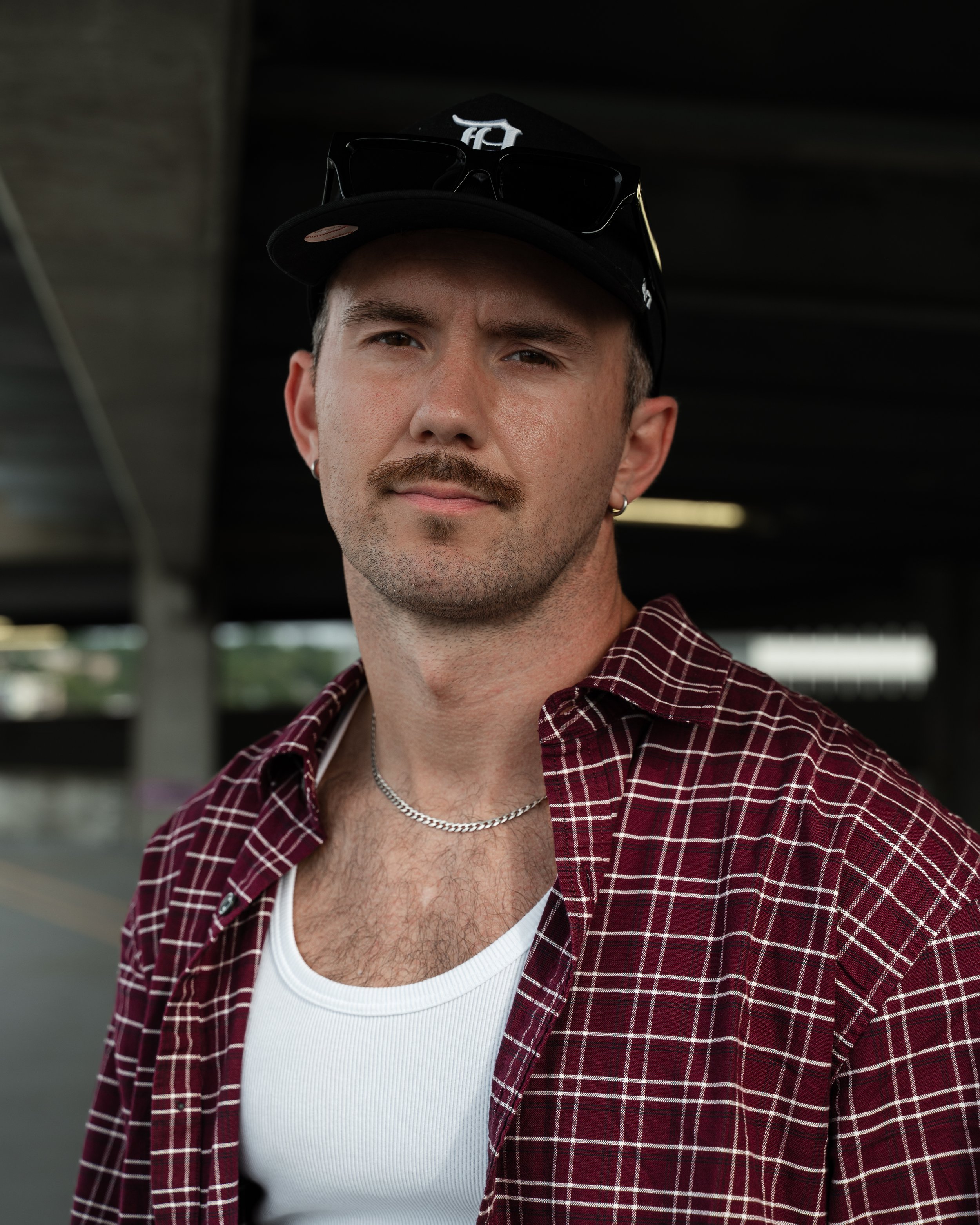 A man with a mustache and beard wearing a black baseball cap with sunglasses on top, a white T-shirt, a red plaid shirt, and a silver chain necklace.