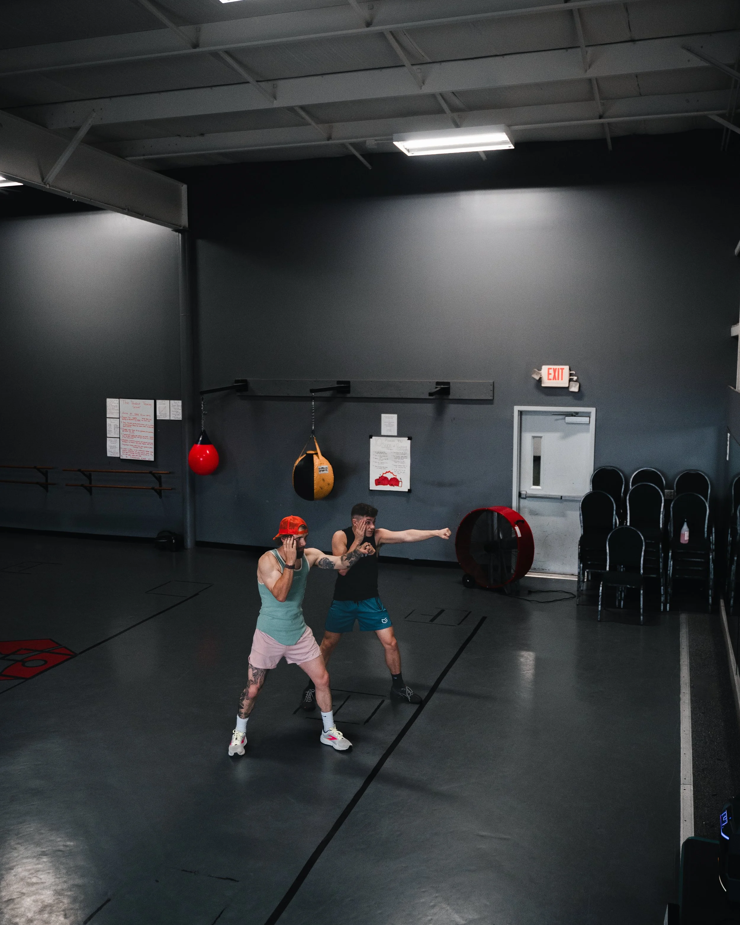Two individuals in a gym or boxing training facility practicing boxing moves. One person is a male with tattoos, wearing a light blue tank top, pink shorts, and white sneakers. The other is a female with a red cap, black tank top, and teal shorts, de