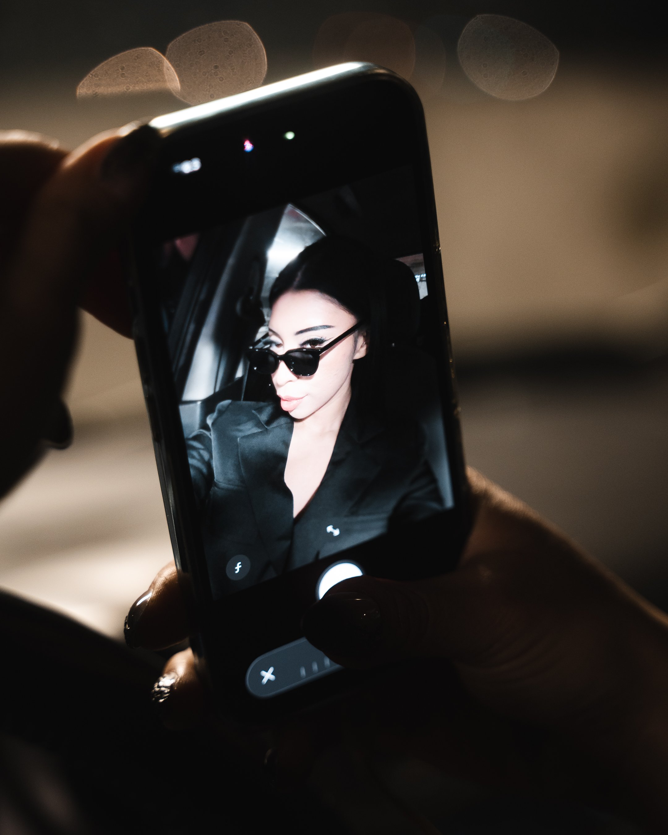 Close-up of a smartphone displaying a woman's selfie with dark sunglasses, held by a hand with painted nails in a dark setting with blurred lights in the background.
