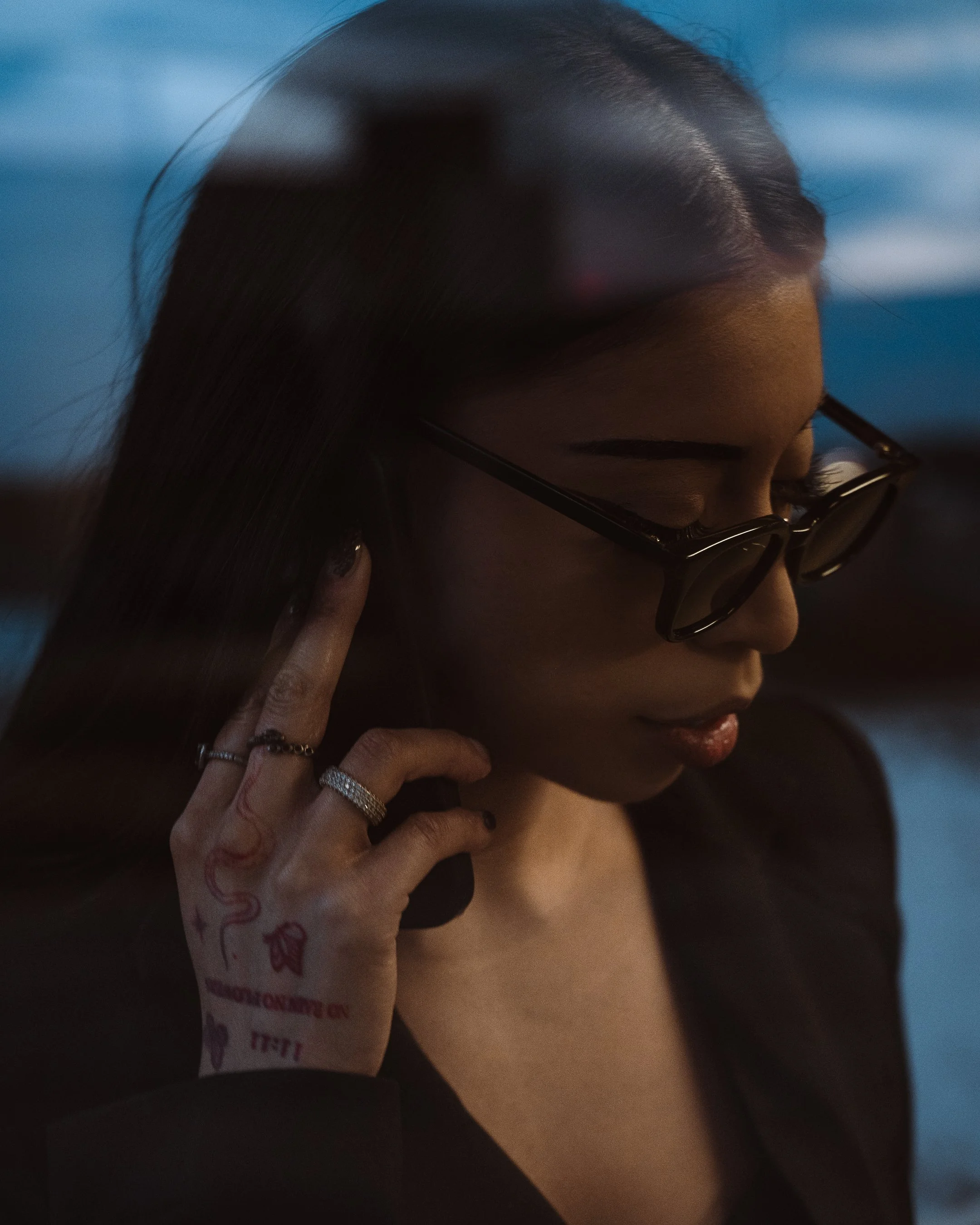 A person with long dark hair and glasses touching their face, wearing jewelry, in a close-up shot.