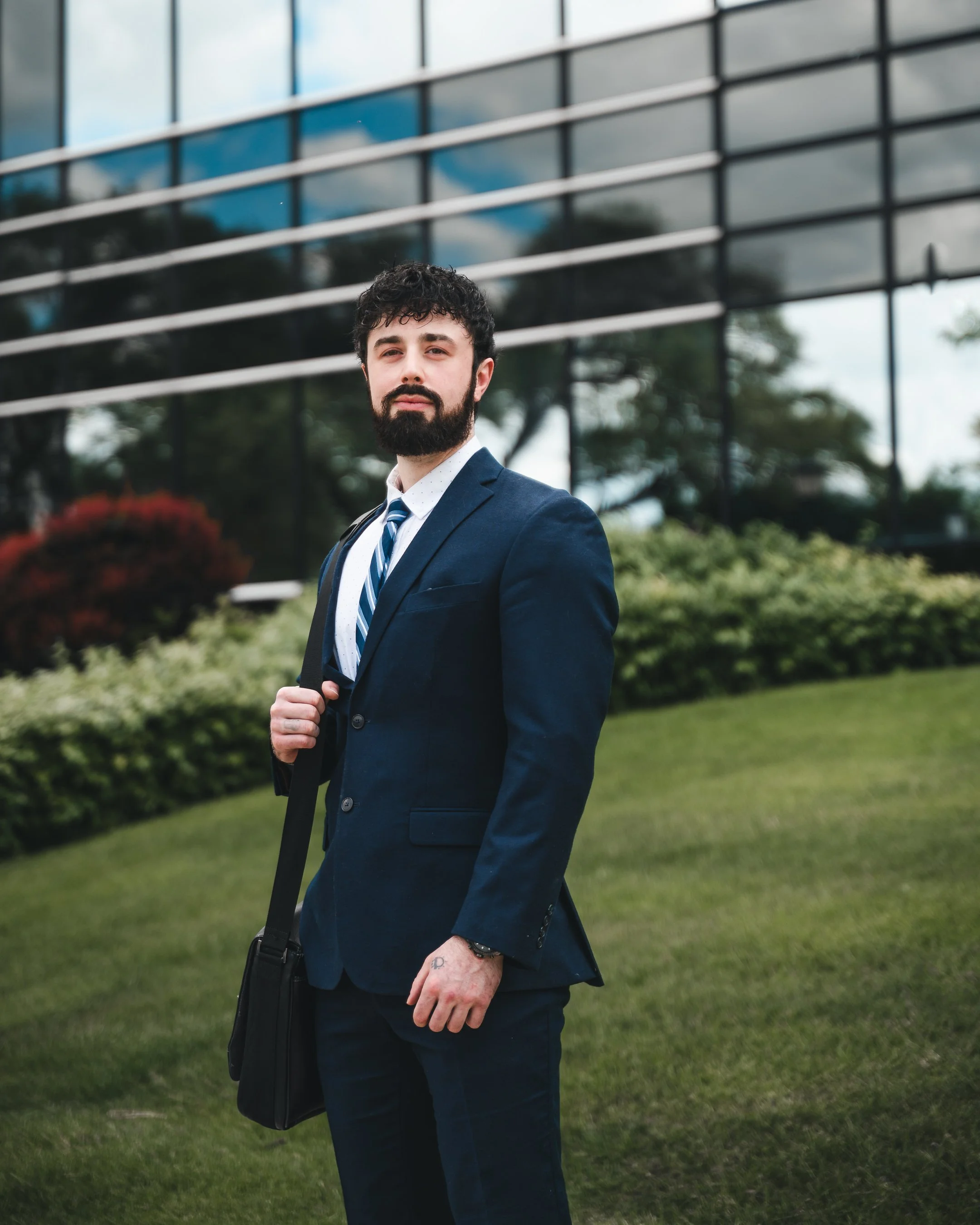 A portrait of a man with dark curly hair and a beard, wearing a dark suit, white shirt, and blue striped tie, standing outdoors in front of a modern glass building and greenery.