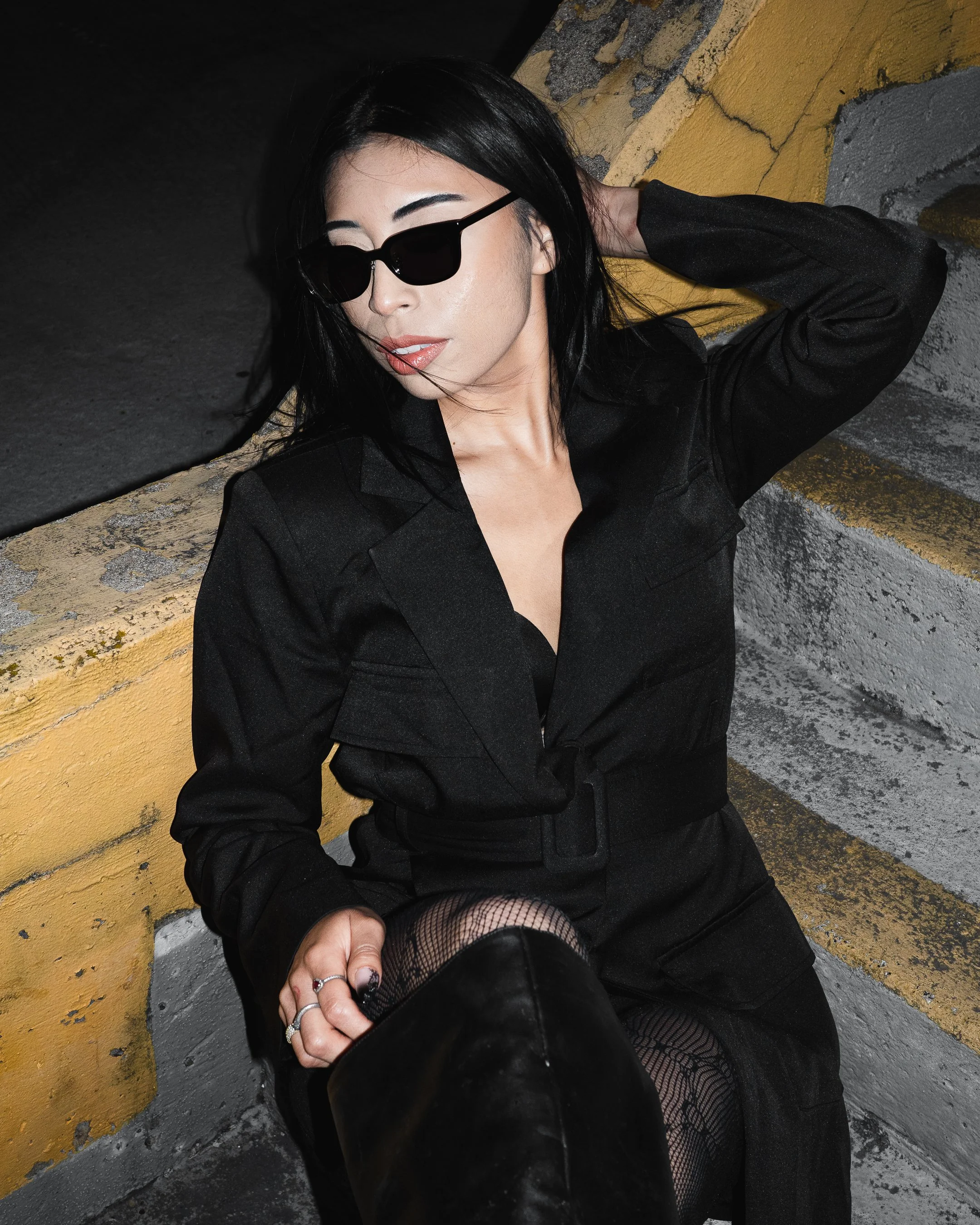 A woman with black hair wearing sunglasses, a black outfit, fishnet stockings, and black shoes, sitting against a yellow and gray textured wall, with a hand near her knee and a ring on her finger.
