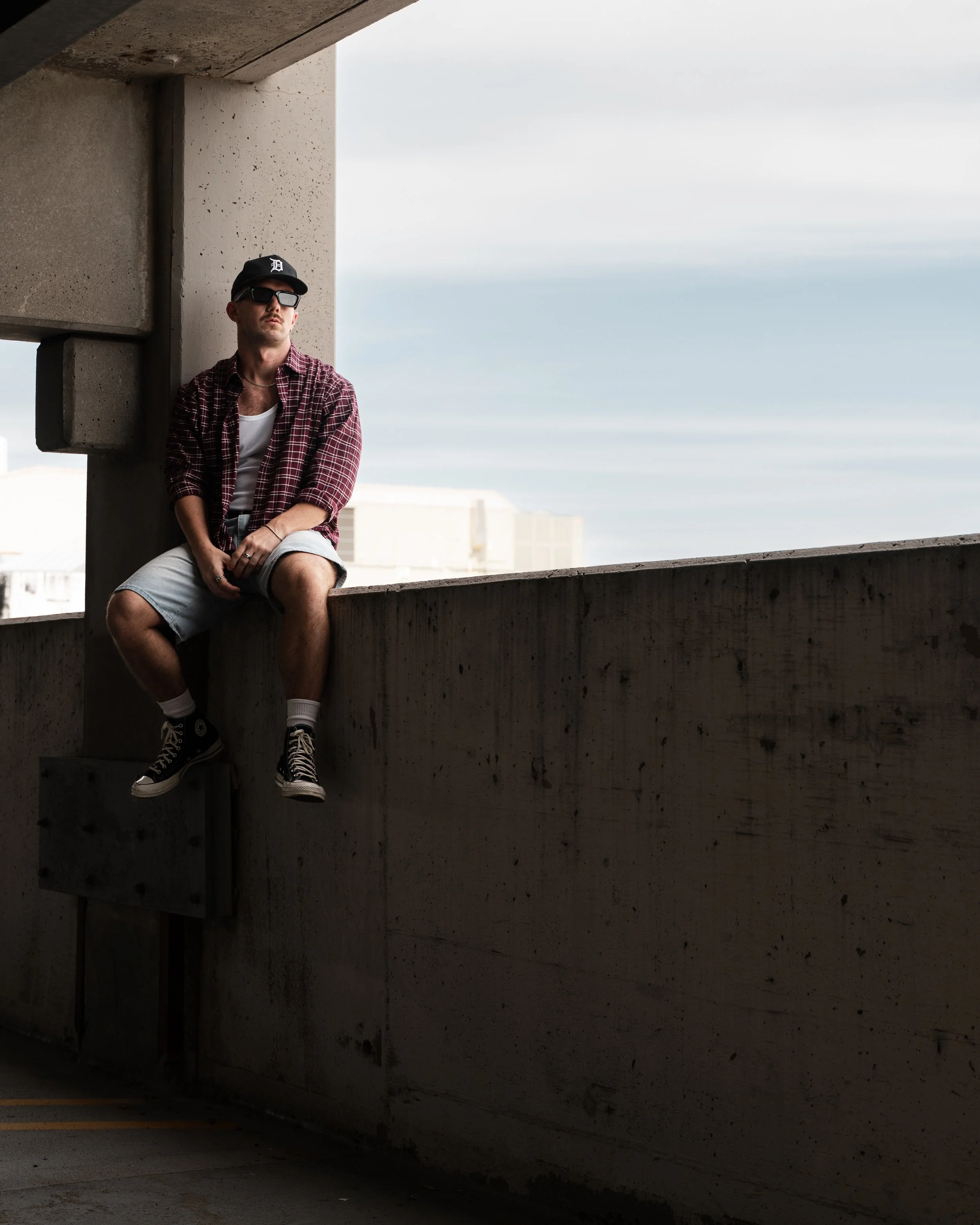 A young man wearing a black baseball cap with a white letter D, sunglasses, a red plaid shirt, a white tank top, beige shorts, and black Converse sneakers, sitting on a concrete ledge under a parking garage with a cloudy sky in the background.