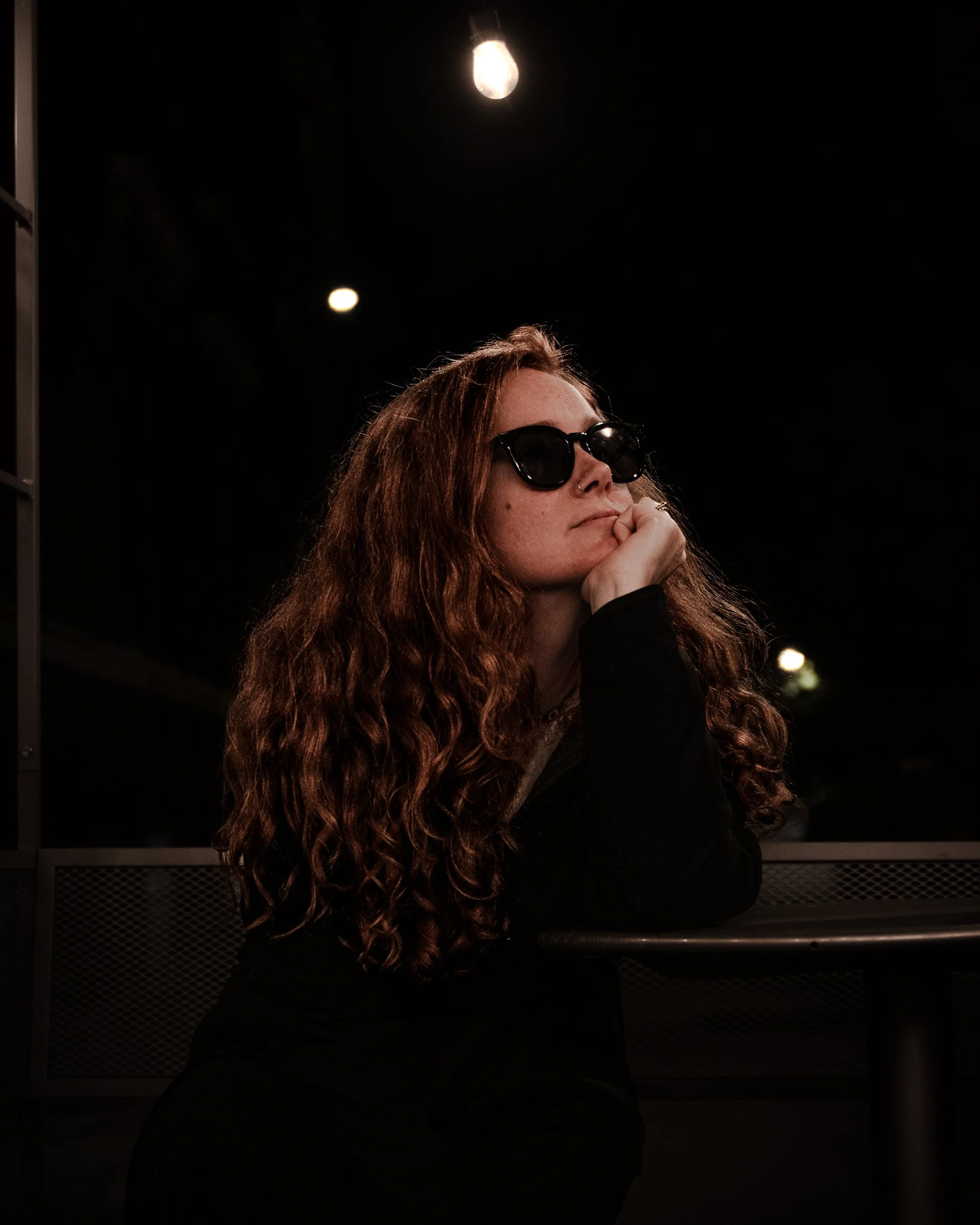 A woman with long red hair wearing sunglasses, resting her chin on her hand, sitting in a dark space with a lit light bulb above her head.