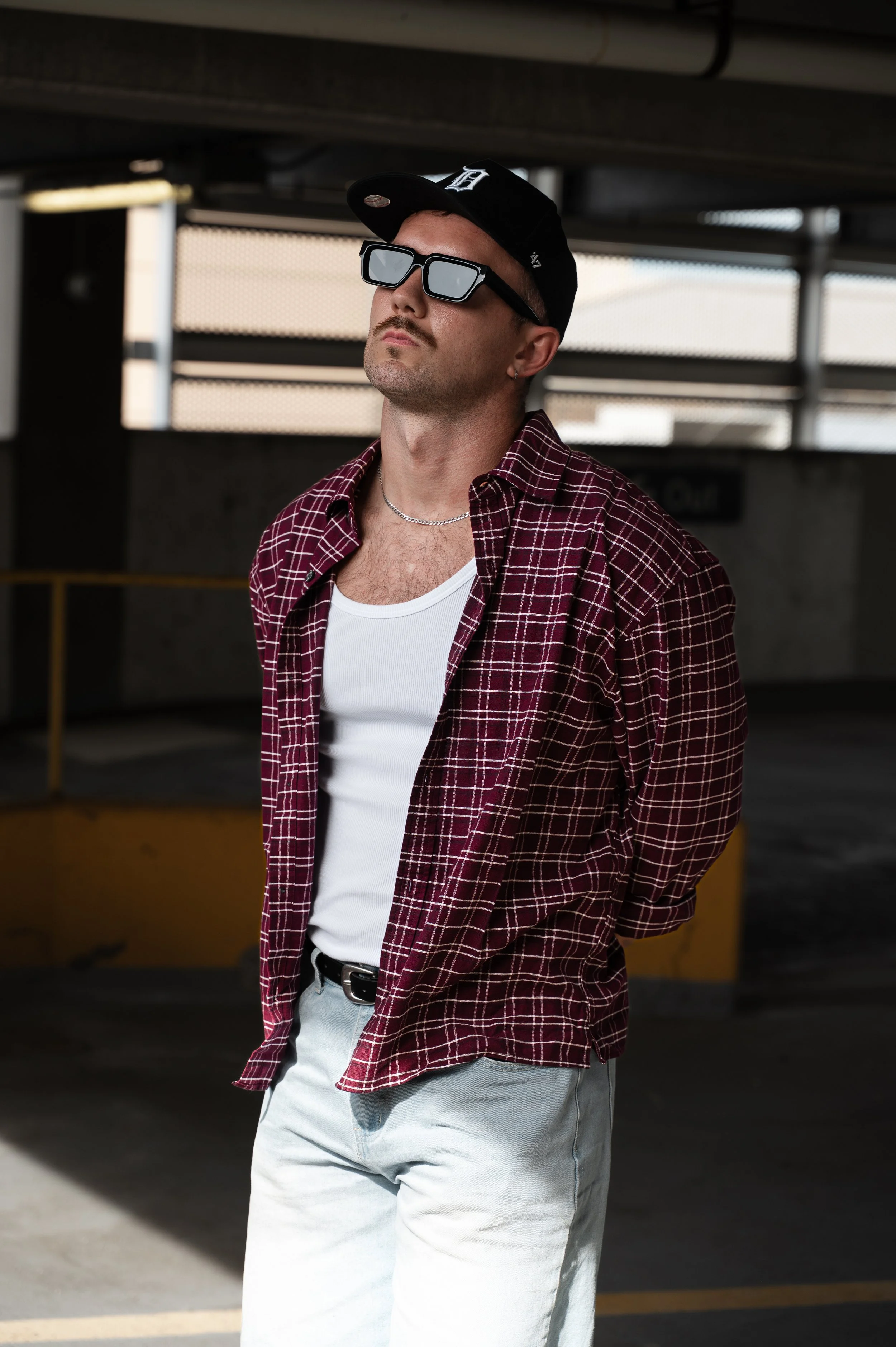 A young man stands in a parking garage wearing a black baseball cap, large black sunglasses, a red and black plaid shirt over a white tank top, and light-colored jeans.