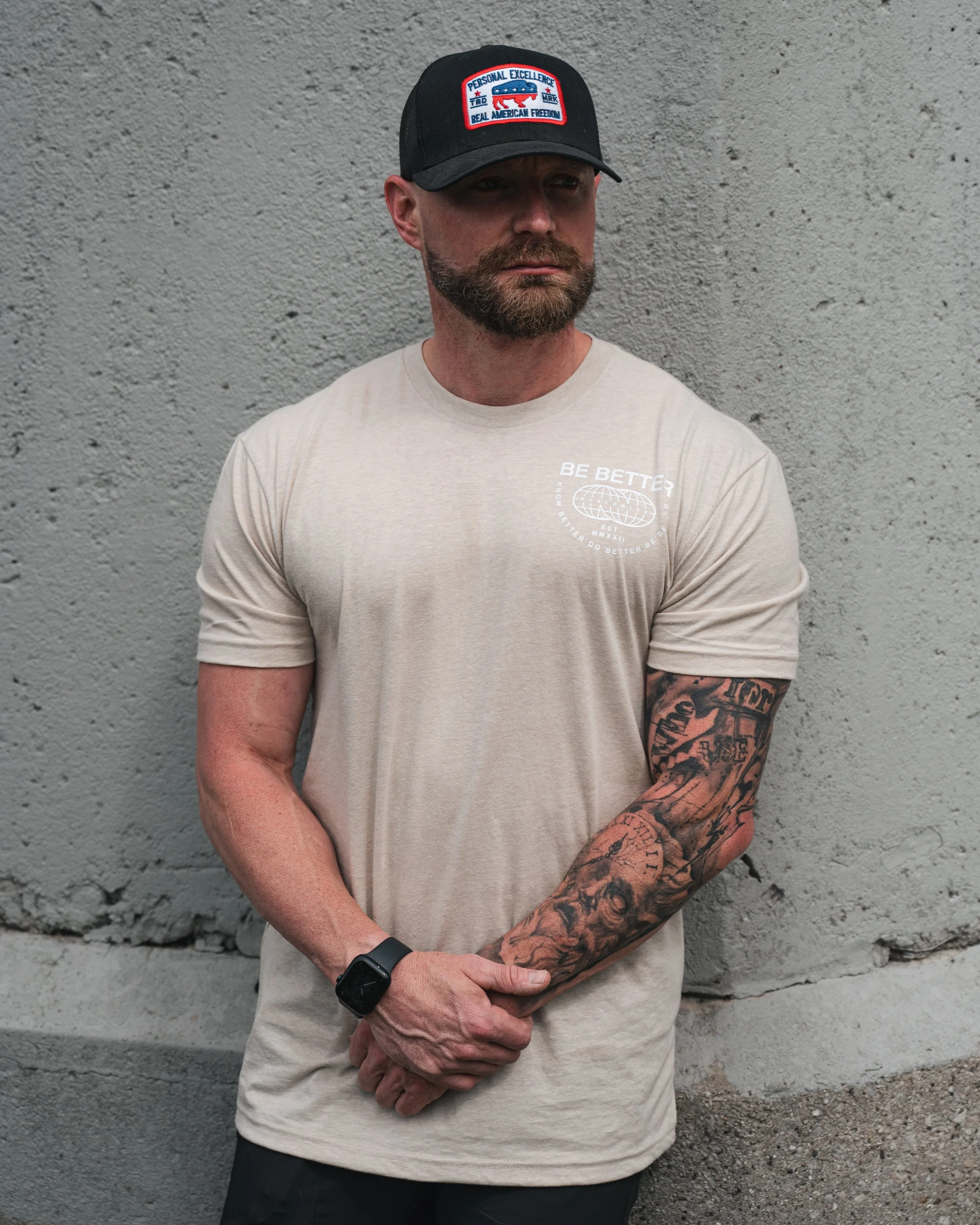 A man with a beard wearing a black cap and a beige t-shirt stands against a gray concrete wall, with his arms crossed.